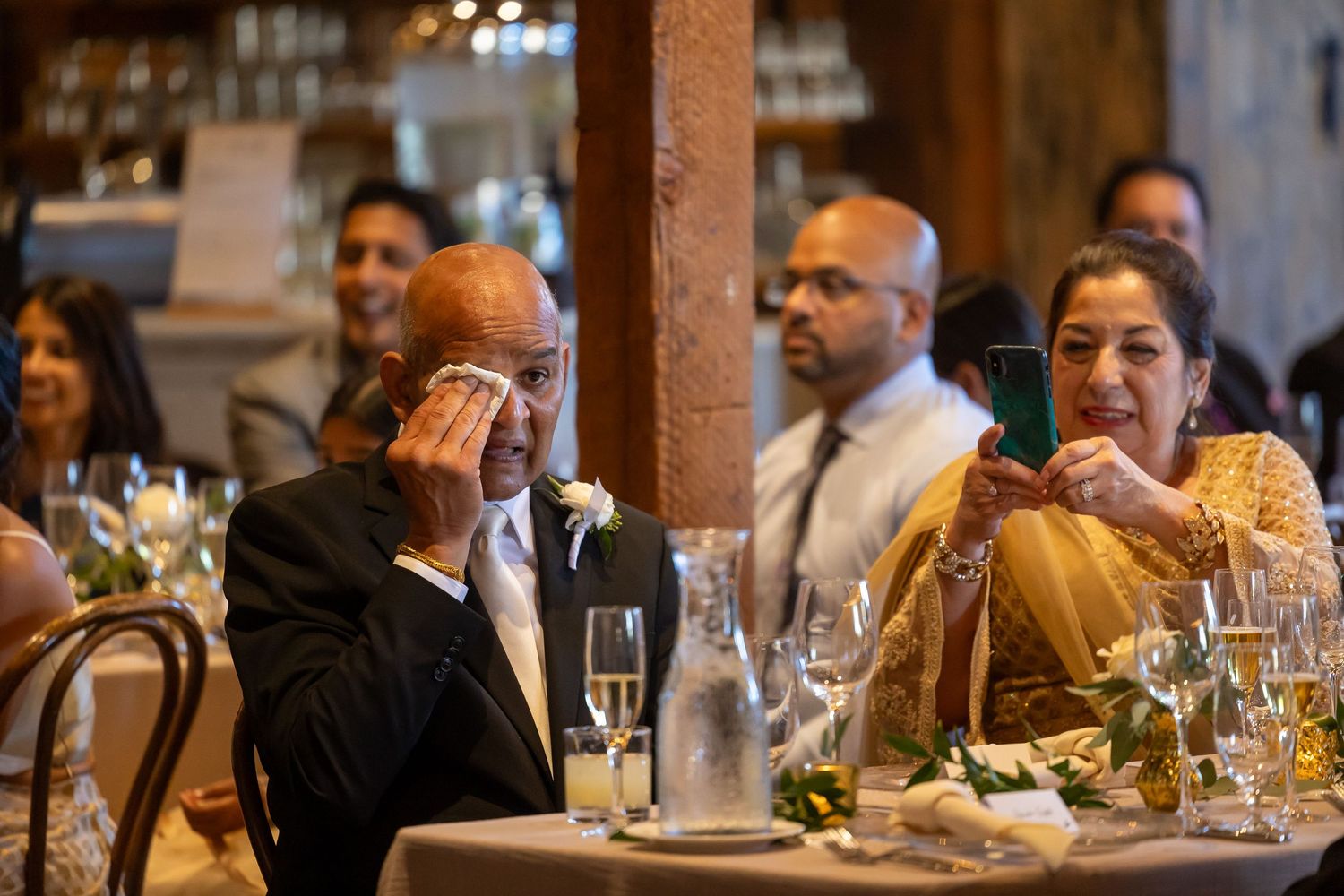 Emotional candid moment captured during wedding reception as guests sit at tables with wine glasses and decor in San Jose.