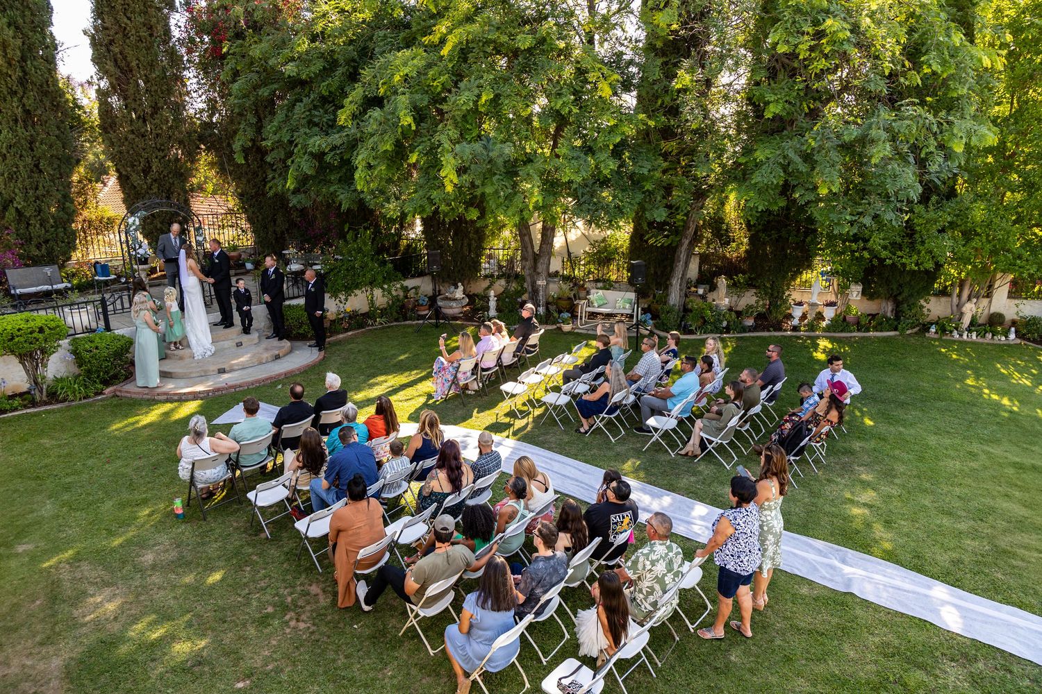 Outdoor wedding ceremony with guests seated on white chairs under trees in a garden setting.