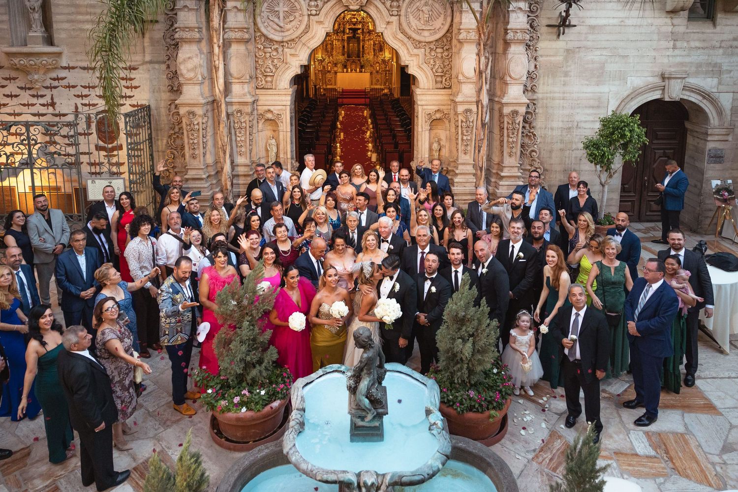 Large wedding party gathered outside ornate church entrance with decorative fountain.