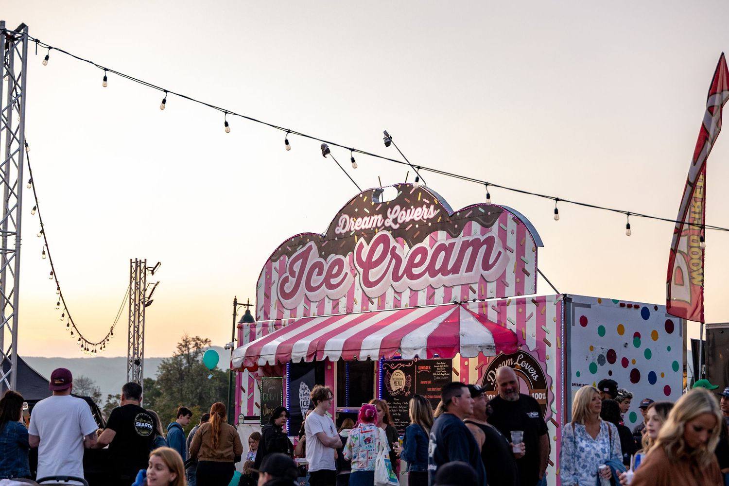 Festive carnival scene at sunset with string lights, colorful food stalls and rides visible in the background.