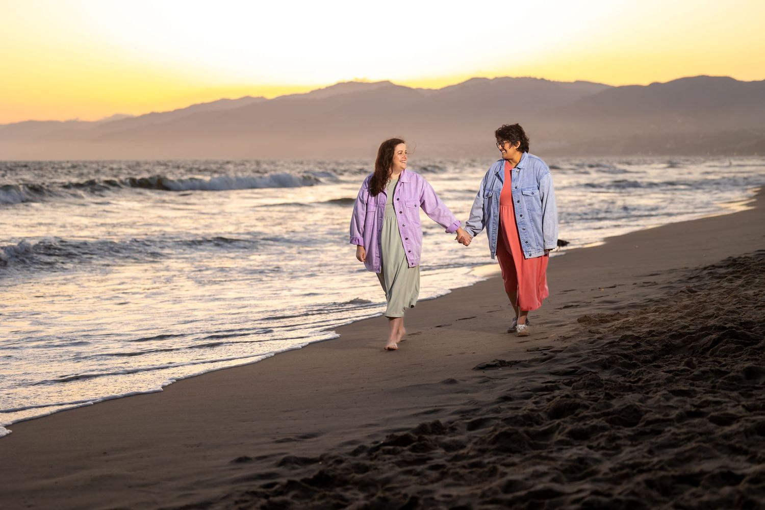 Couple walking hand in hand on beach at sunset wearing colorful outfits.