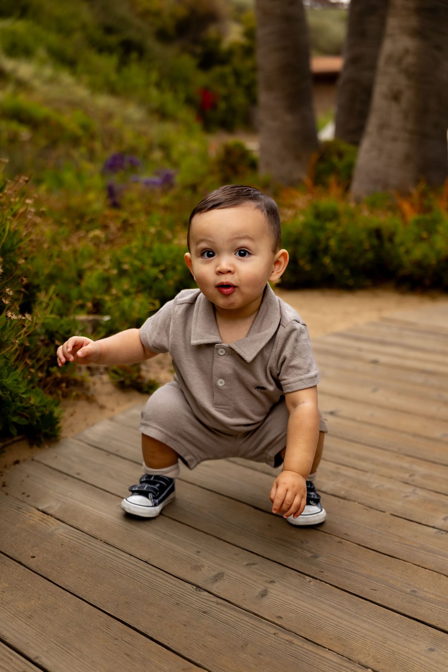 Baby in grey polo outfit exploring wooden deck outdoors.