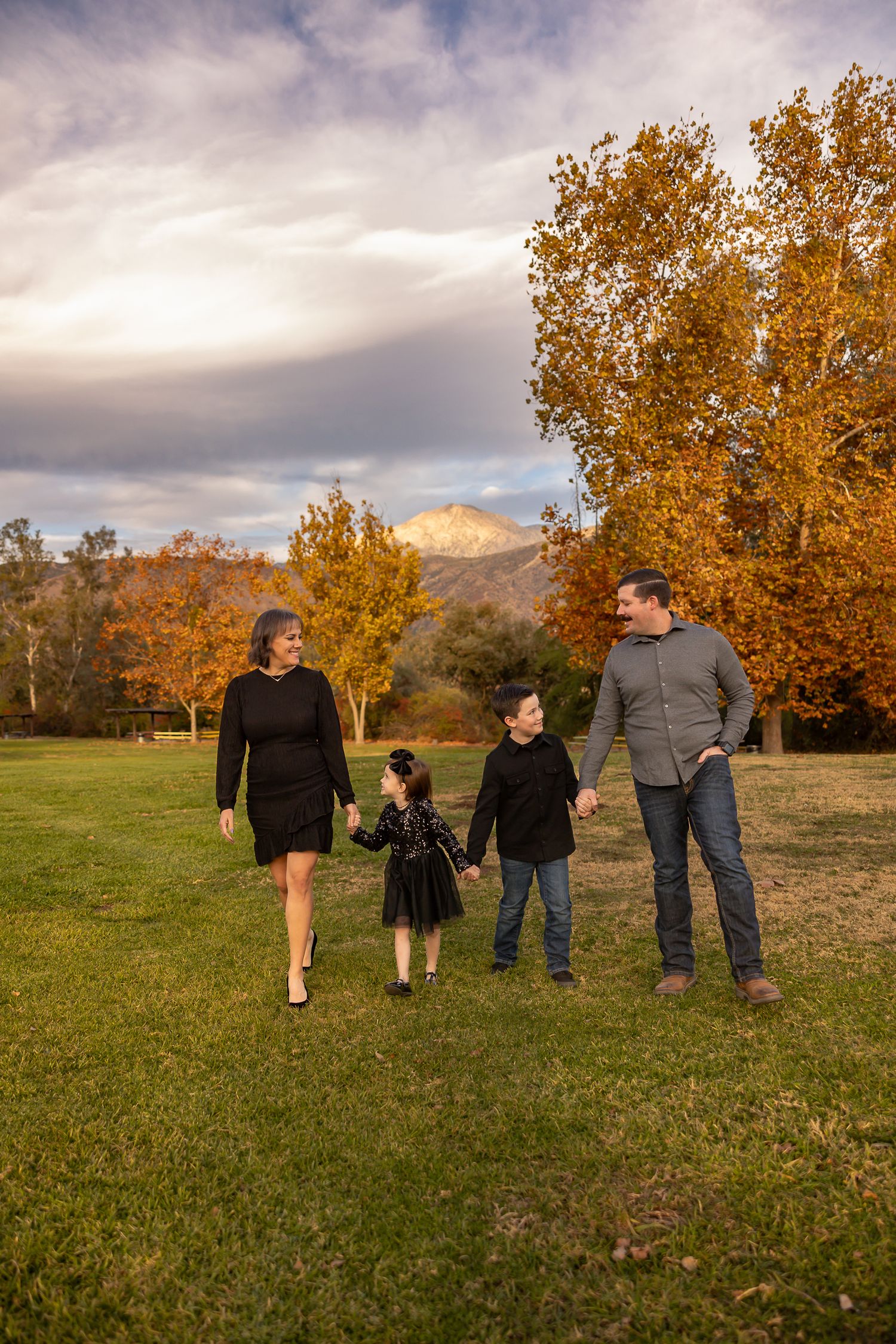 A family walks together across a grassy field with golden autumn trees behind them.