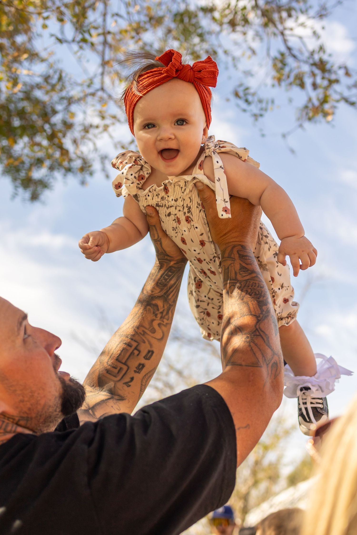 Baby wearing red bow and floral romper being lifted against blue sky.