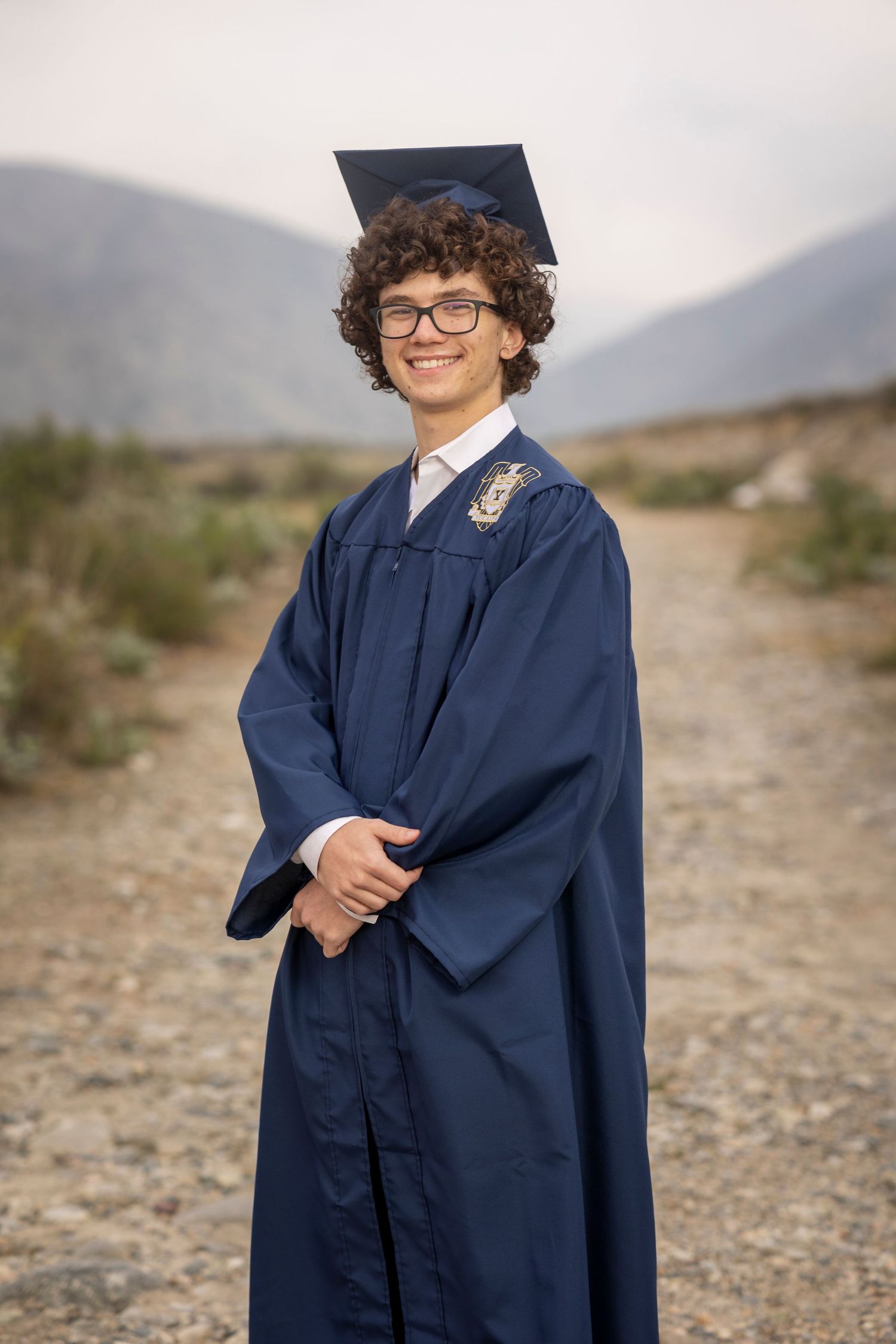 Graduate in navy blue cap and gown standing on desert path with mountains in background in Yucaipa.