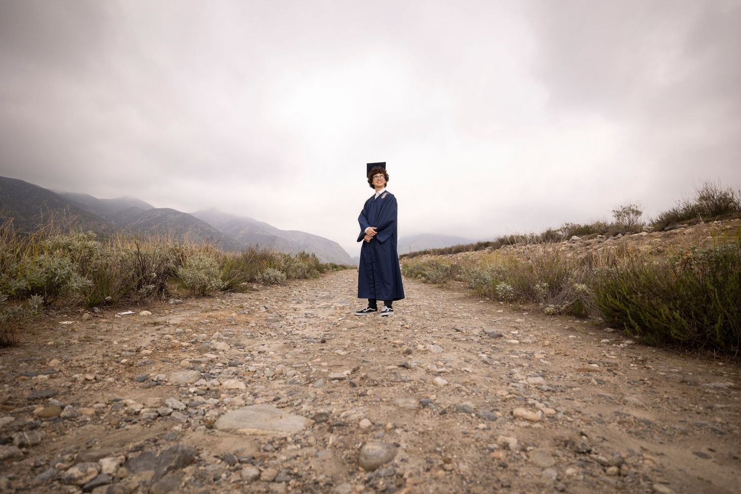 Graduate in navy gown standing on dirt path with misty mountains in background in Yucaipa.