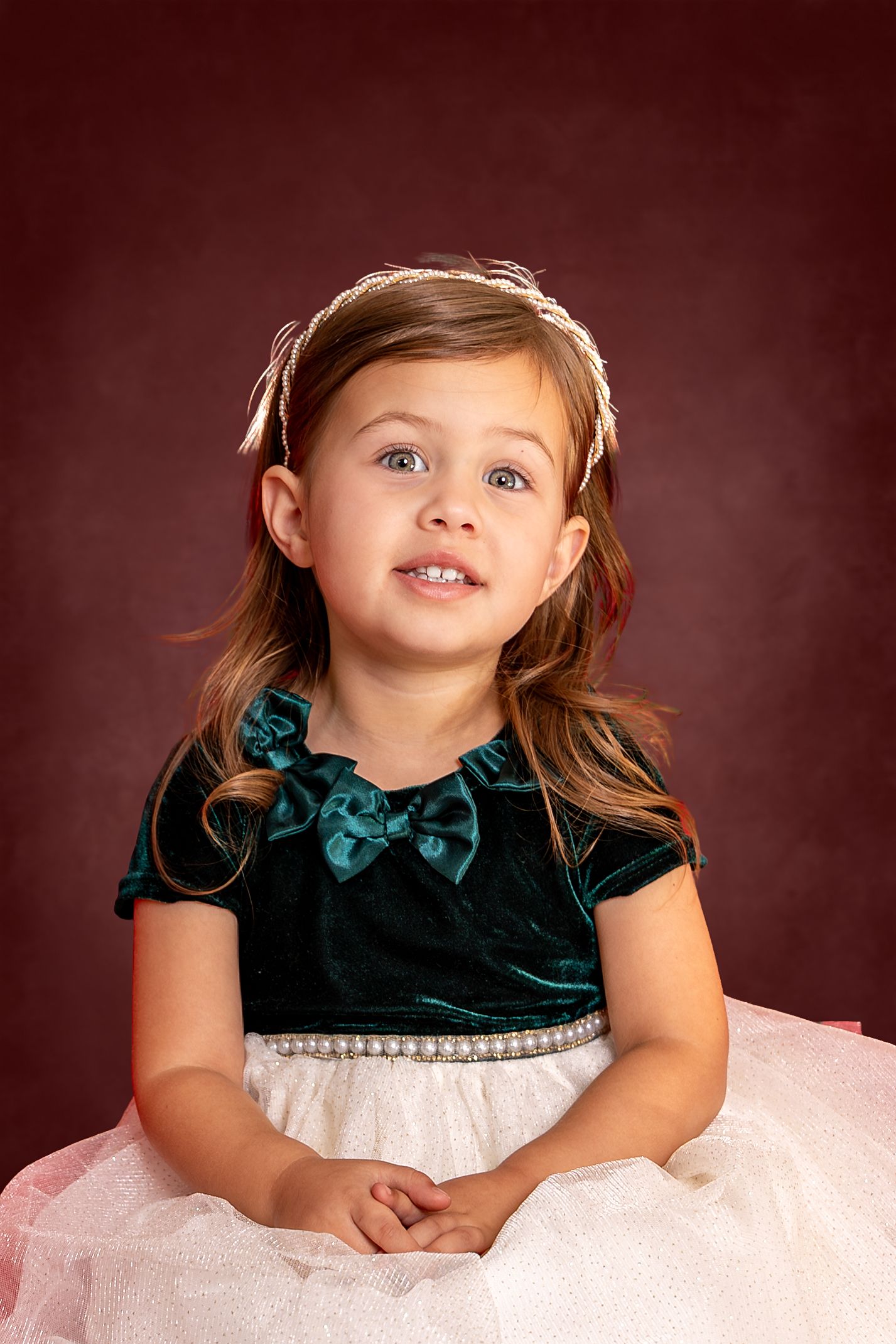 Young girl in teal velvet dress with white tulle skirt and pearl belt, smiling against dark red background.