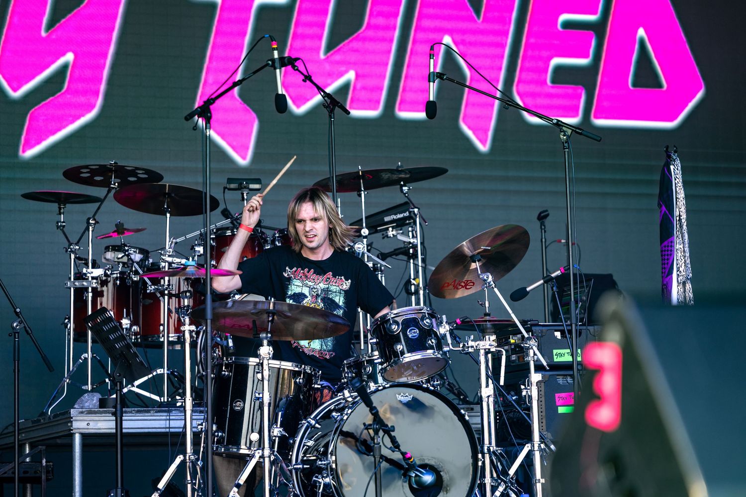 Drummer performing on stage with pink neon signage behind a professional drum kit.