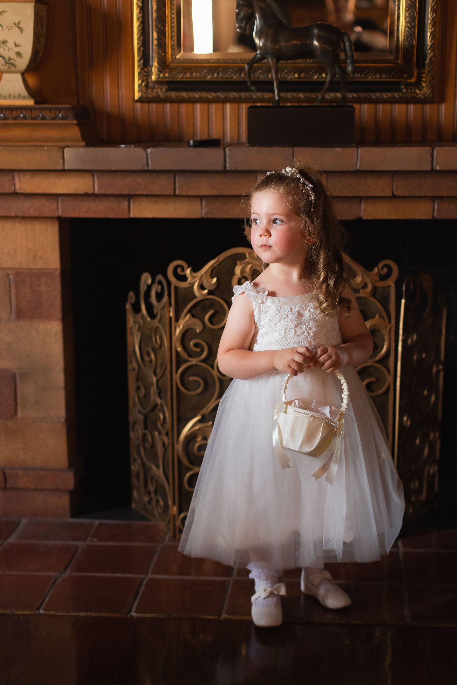Flower girl in white tulle dress standing by brick fireplace holding basket.