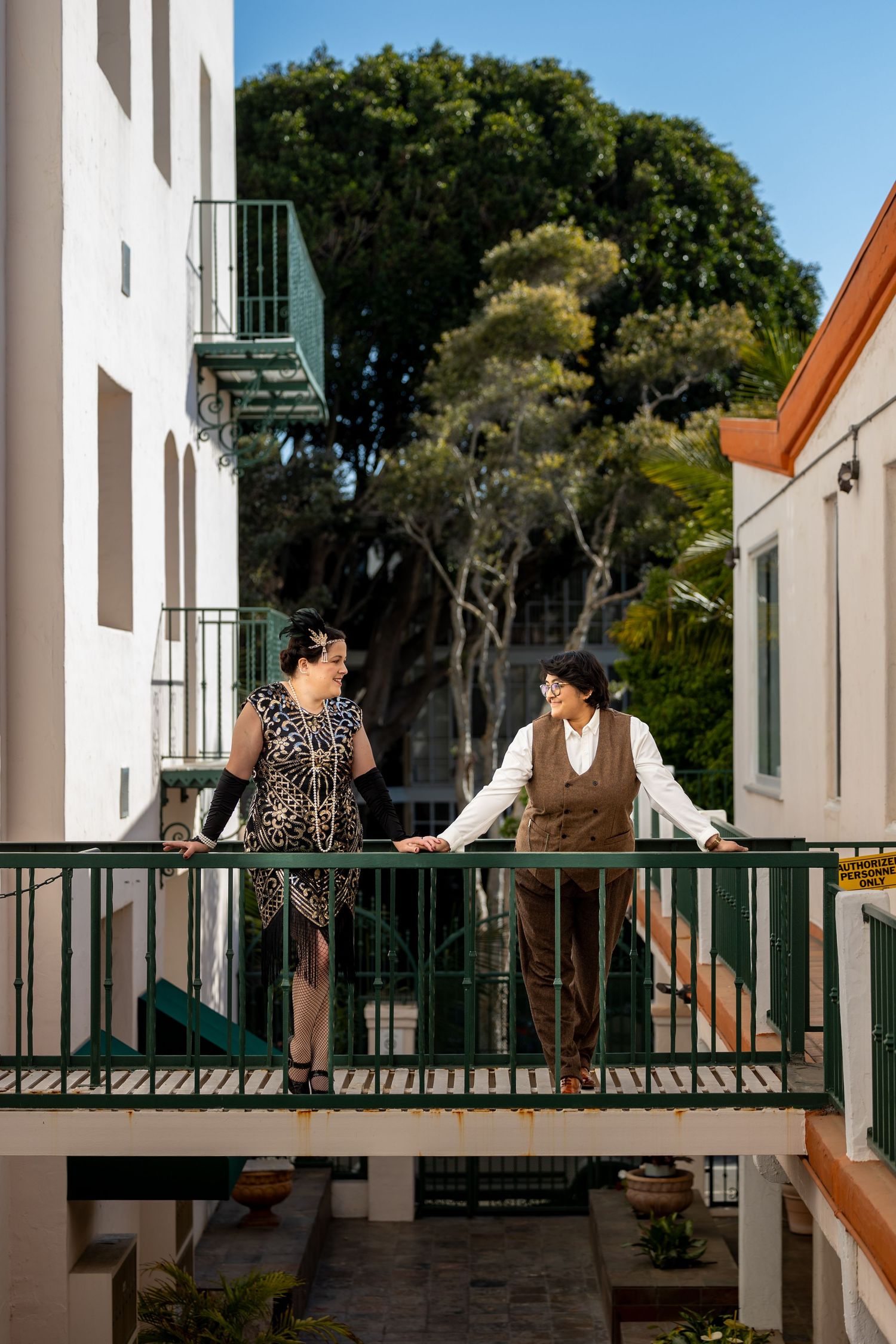 A stylishly dressed couple stands on a green balcony between white buildings.