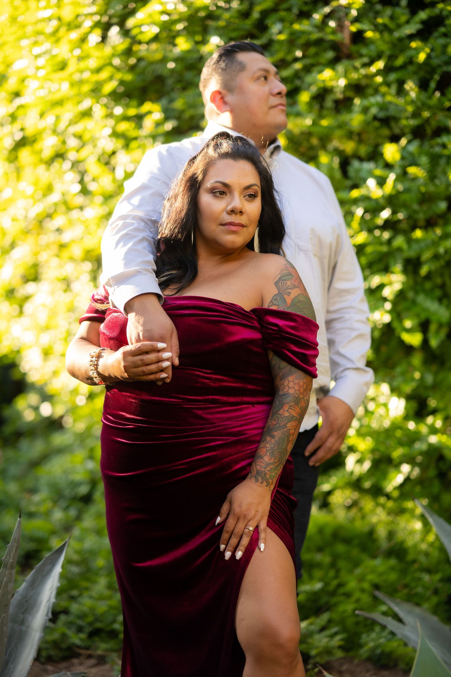 A romantic couple poses in a garden setting with woman wearing burgundy velvet dress with leg slit.