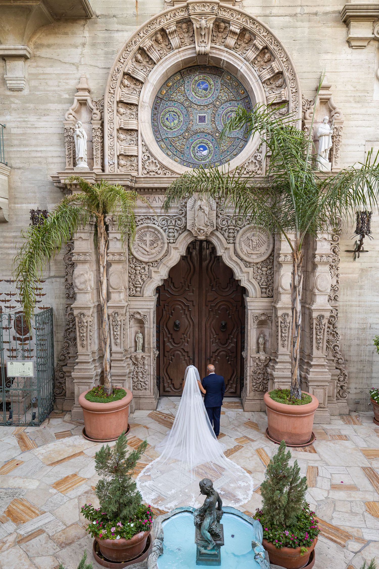 A couple stands before an ornate church entrance with detailed stone carvings, a circular stained glass window, and palm trees.