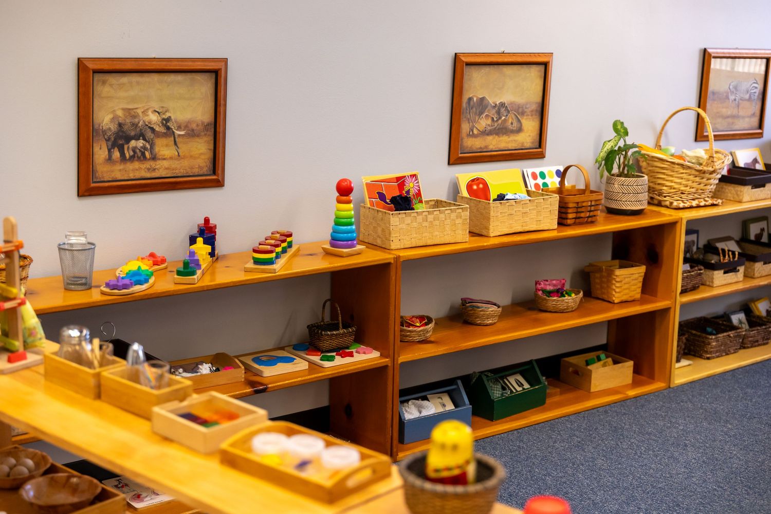 Montessori shelves lined with colorful learning materials, wooden toys, and framed artwork on blue carpeted classroom floor.