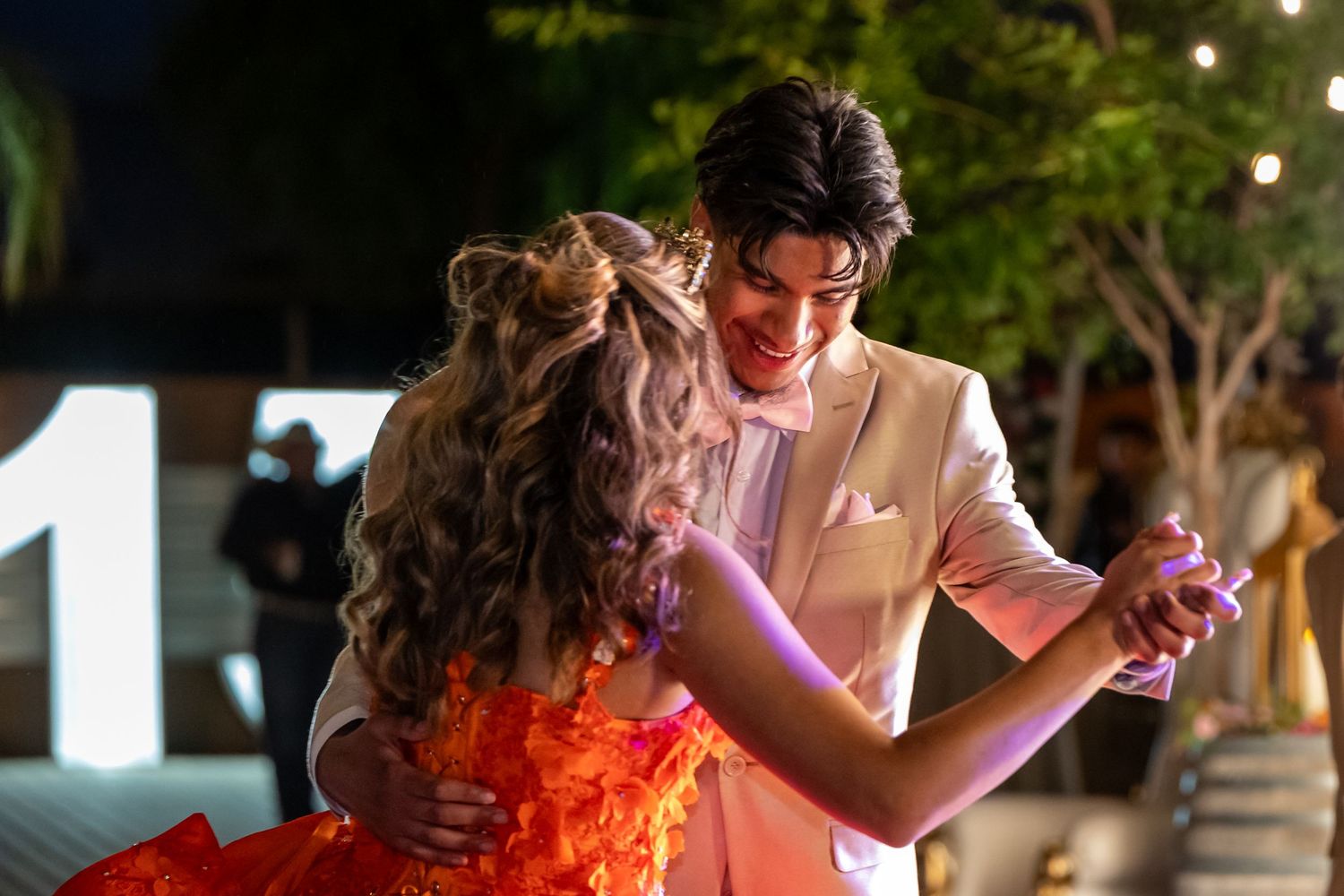 Couple dancing together at night under string lights with one wearing a coral dress.