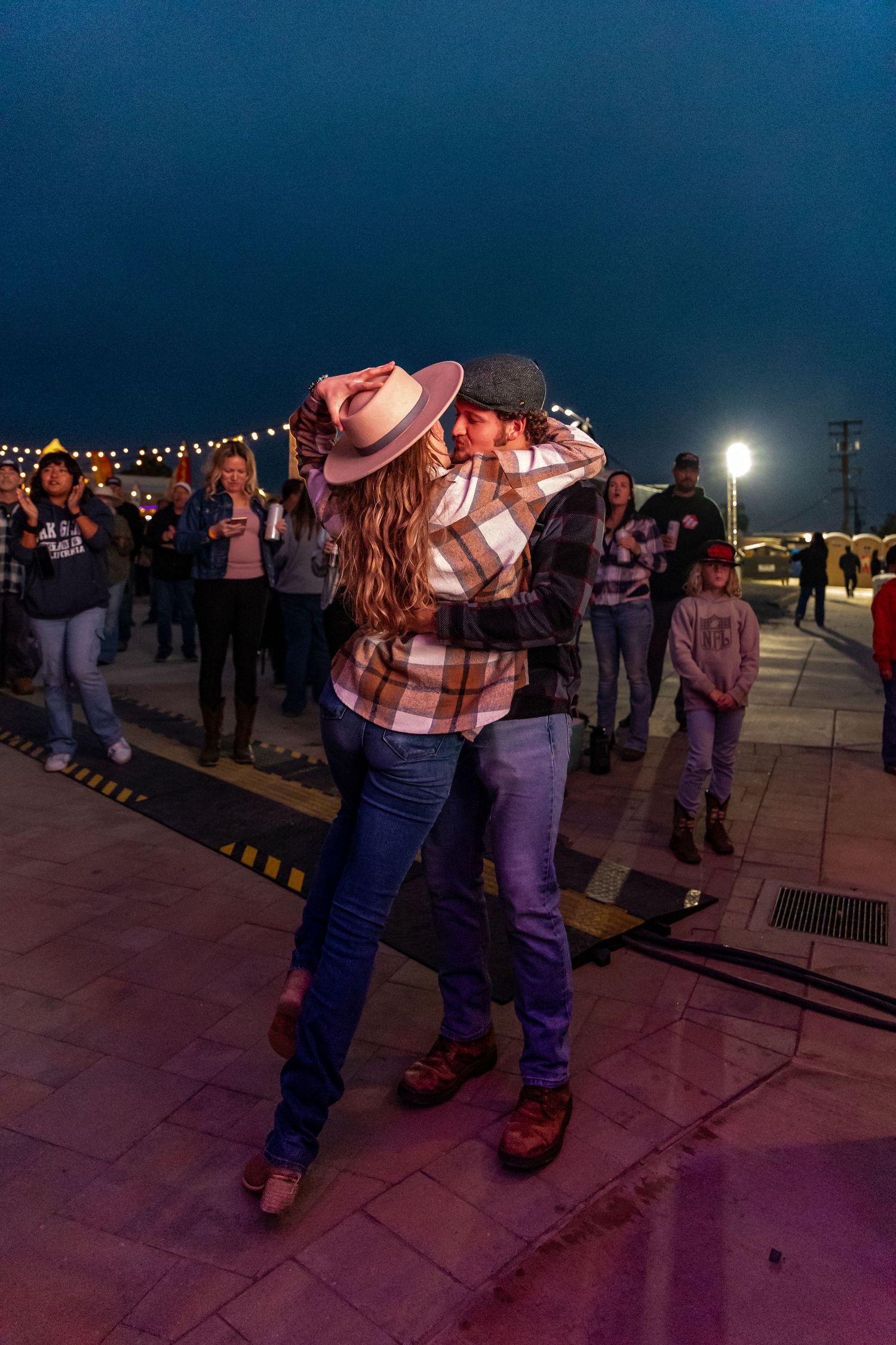 Couple dancing together at night under string lights wearing plaid shirts and cowboy hats.