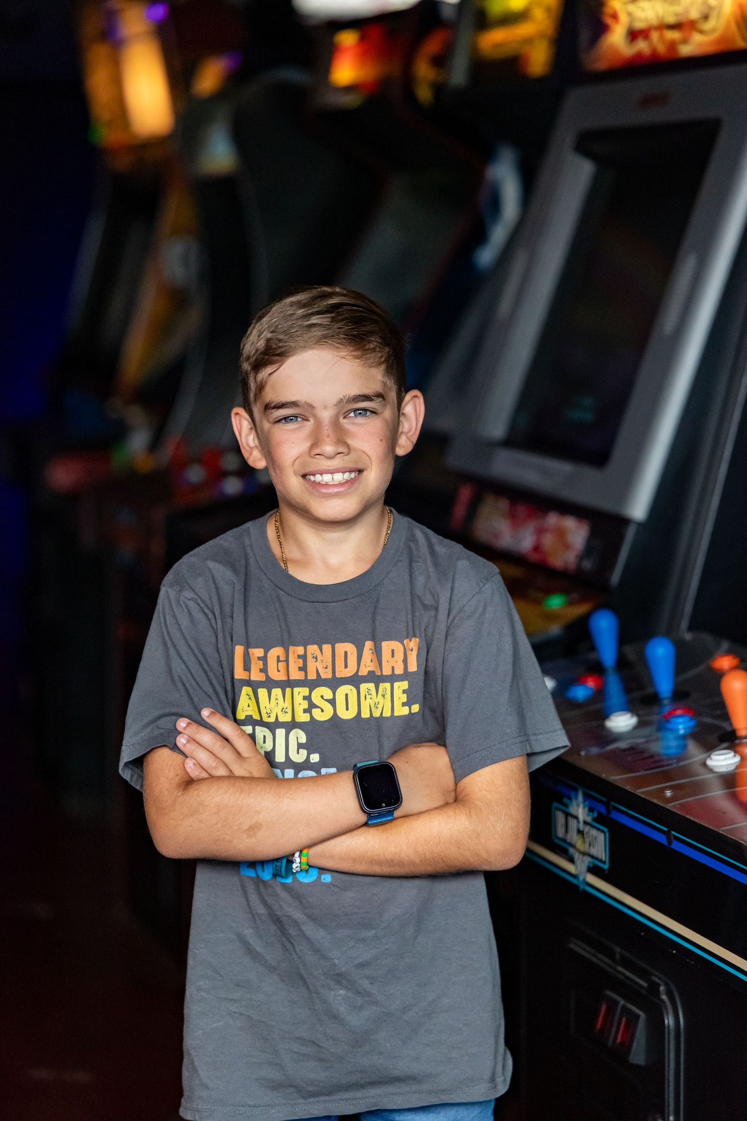 Young person in grey t-shirt stands confidently in front of arcade game machines.