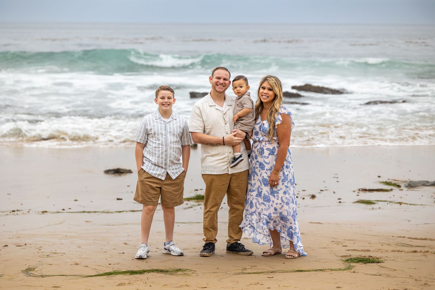 A family poses together on a scenic beach with crashing waves and rocky coastline in the background.