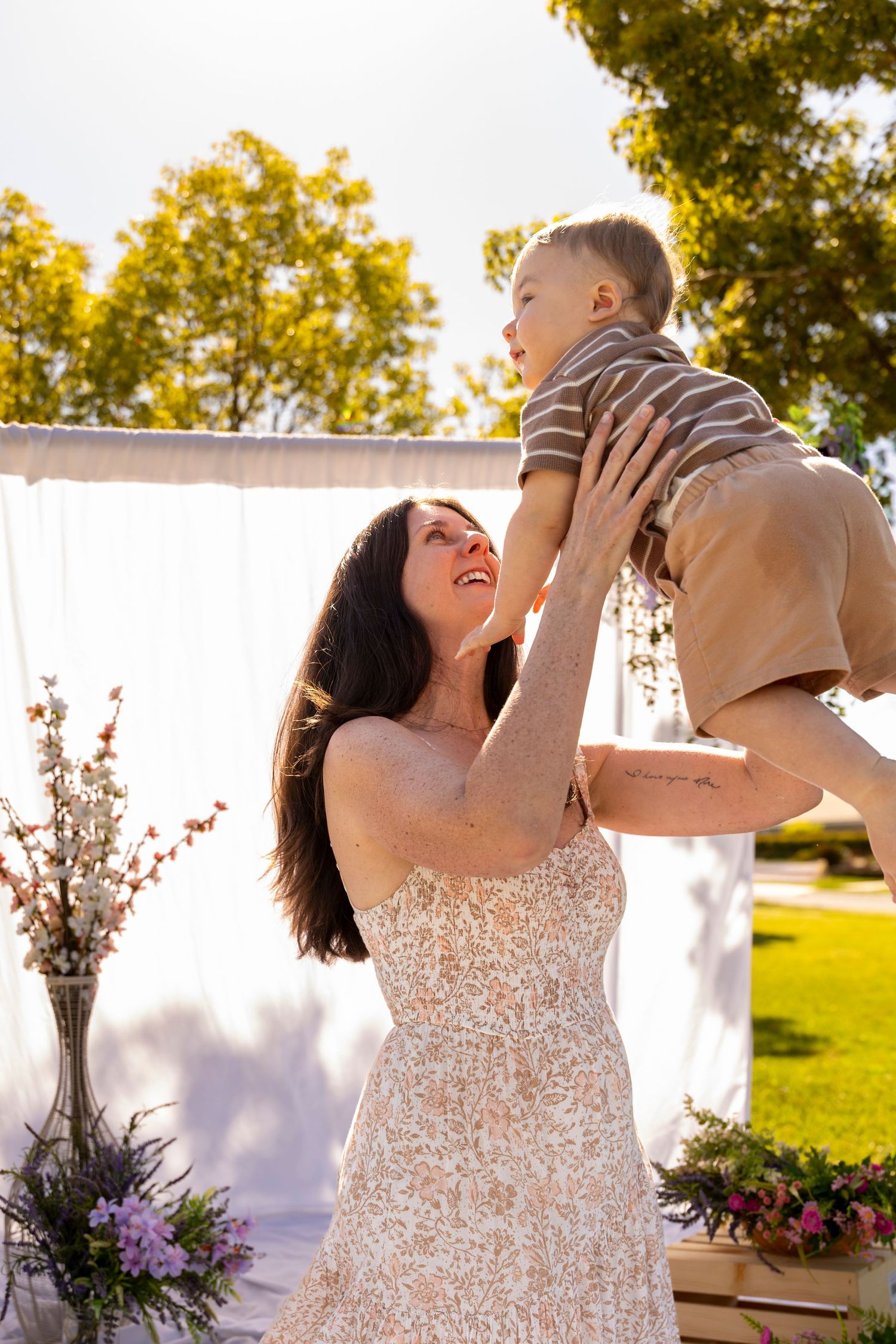 A joyful woman in a floral dress lifts a smiling toddler boy outdoors near a decorated white arch.