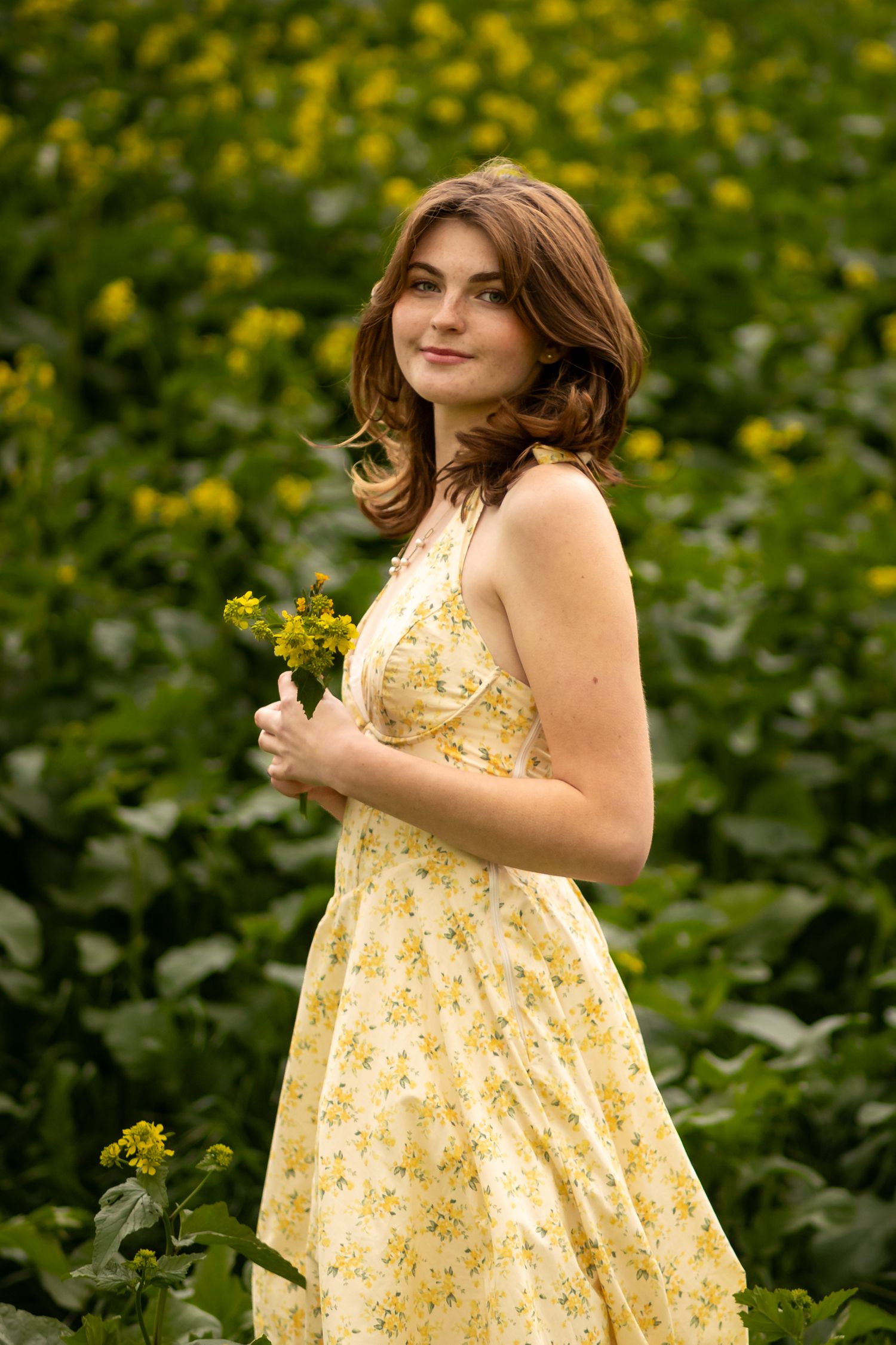 Senior in yellow floral sundress poses in a garden filled with blooming yellow flowers on a sunny day in Redlands.