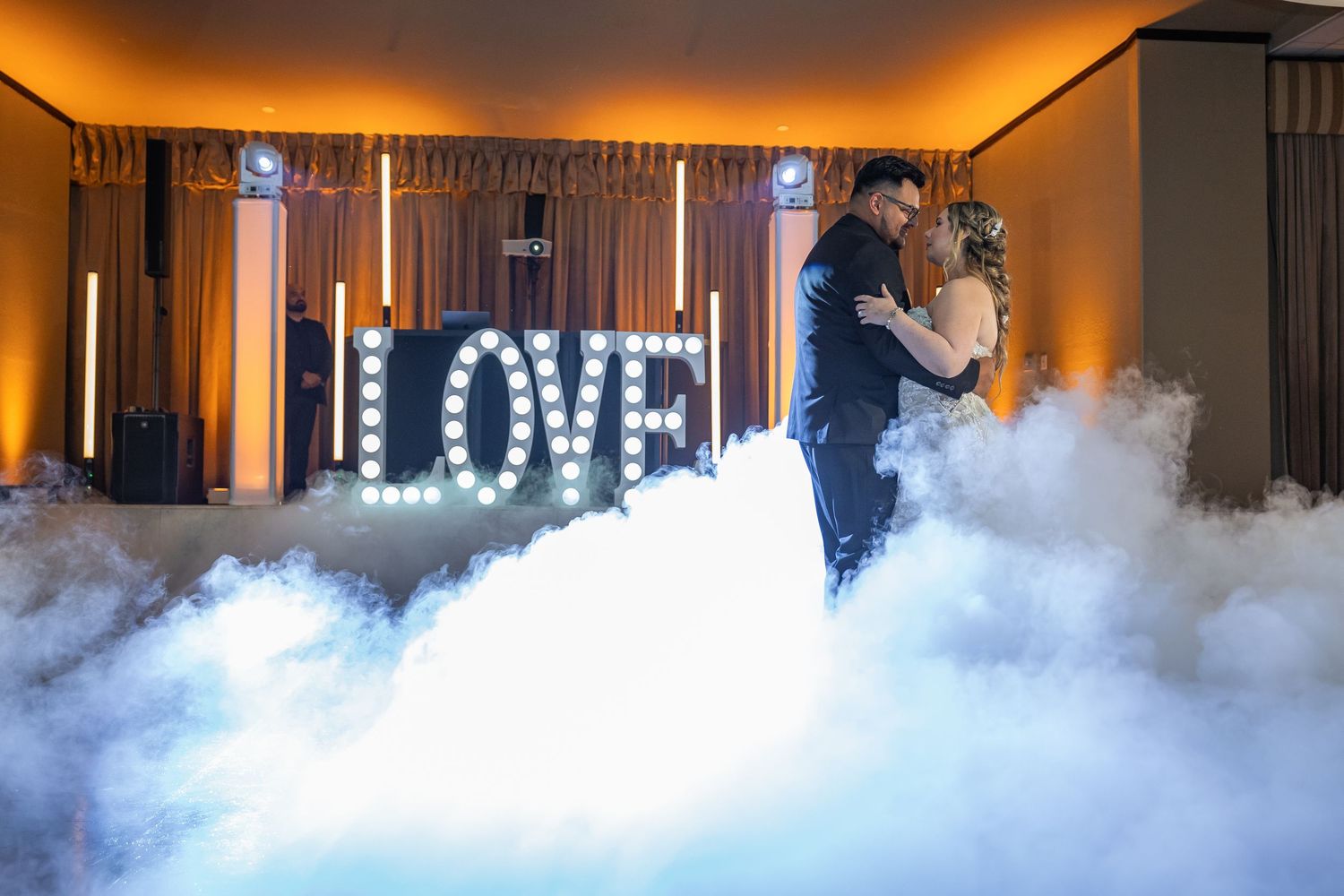 Couple dancing on a cloud of fog with illuminated LOVE sign glowing in the background at a wedding reception.