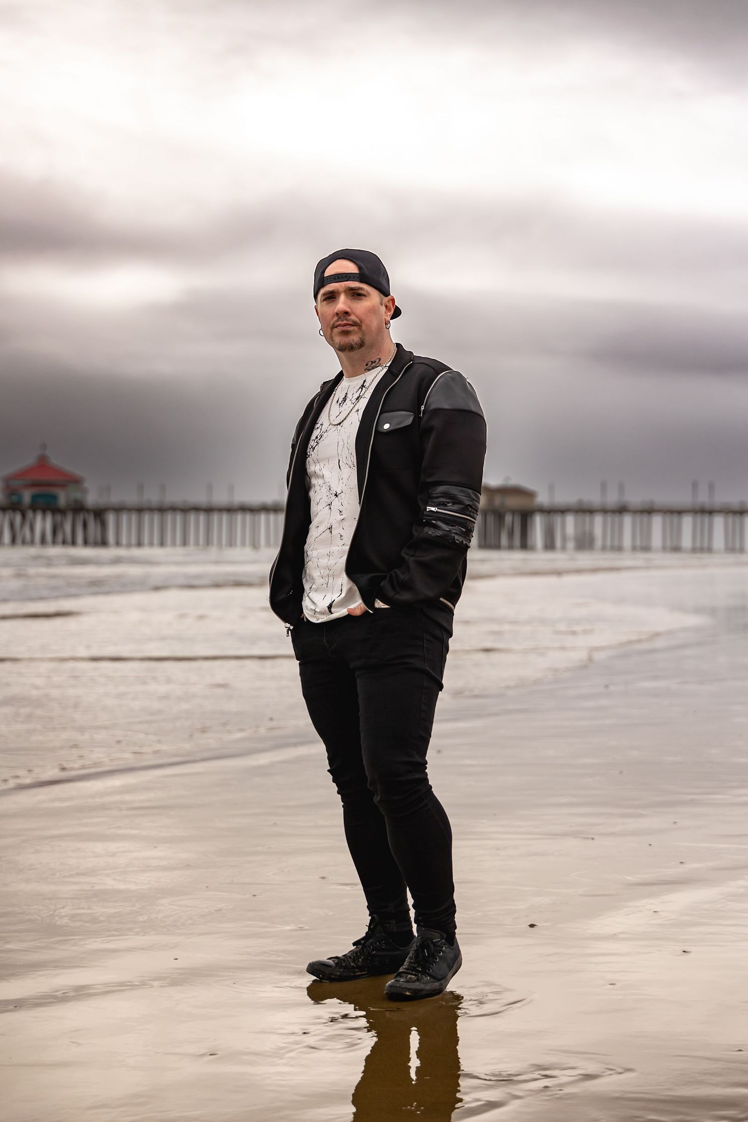 Person in black jacket standing on wet beach with pier in background in Huntington Beach.
