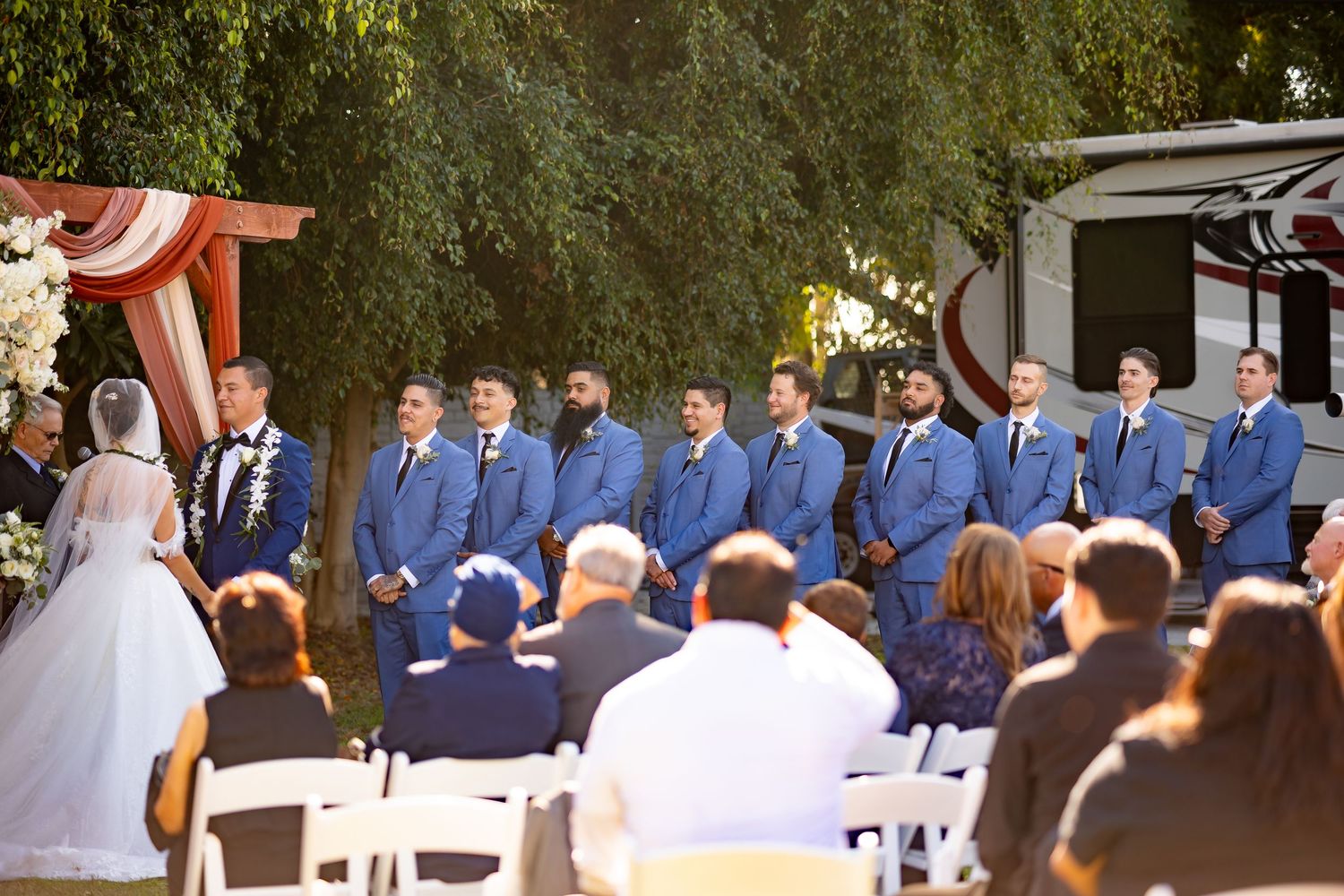 A wedding ceremony in progress with groomsmen in blue suits standing under trees.