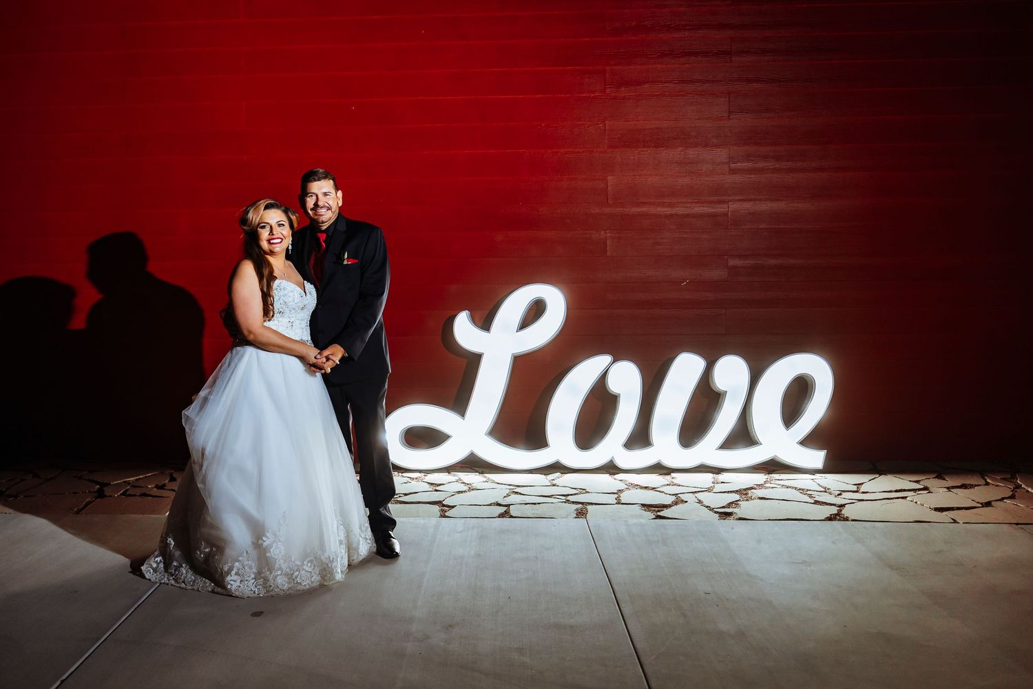 Newlyweds pose against red wall with illuminated LOVE sign at wedding reception.