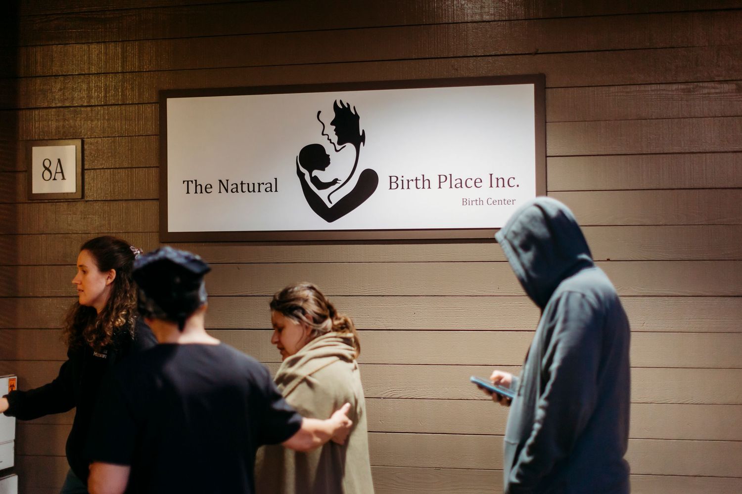 Hospital hallway sign showing 'The Natural Birth Place Inc.' with people waiting.