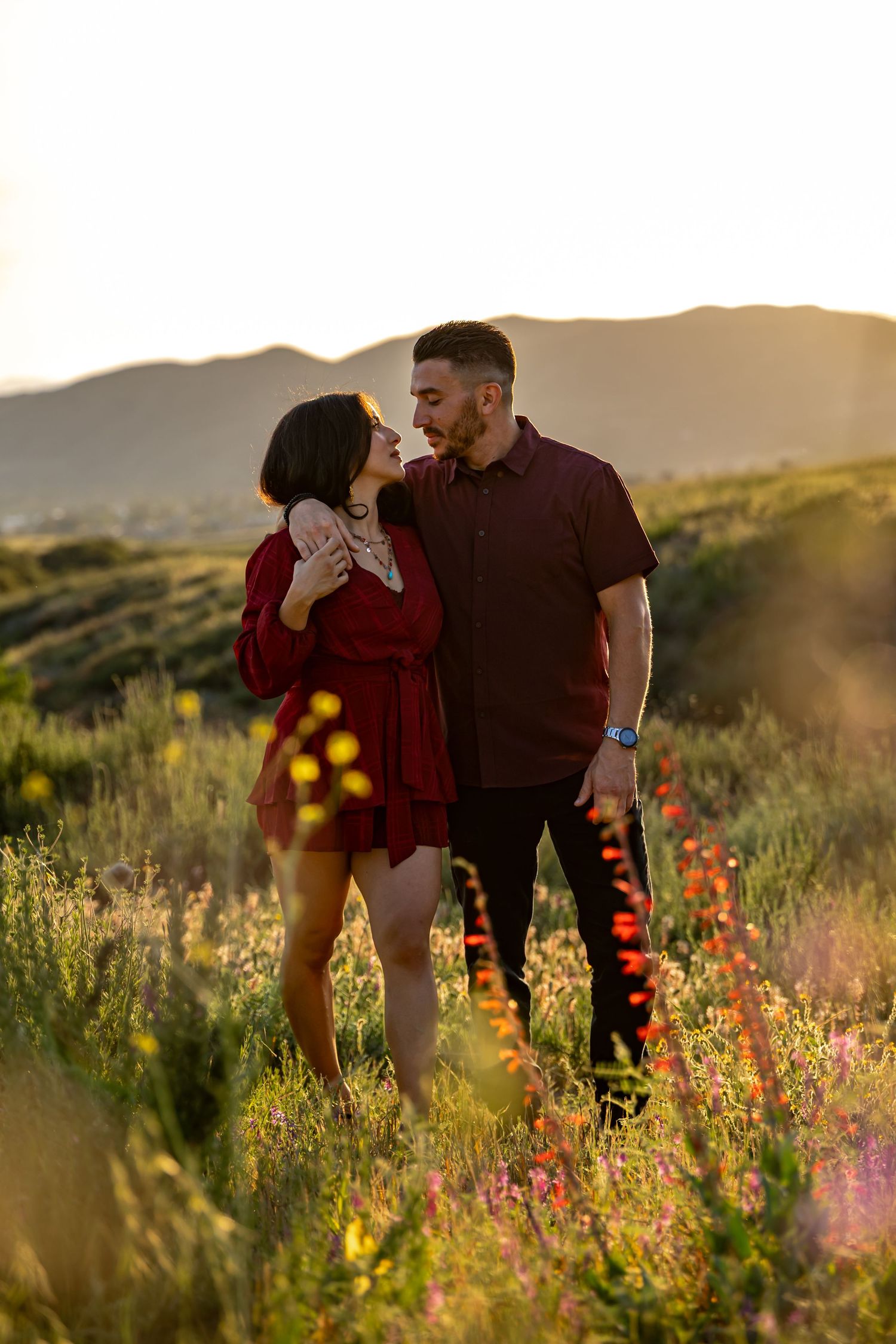 Couple in red and purple embracing in a wildflower field at sunset with mountains in background.