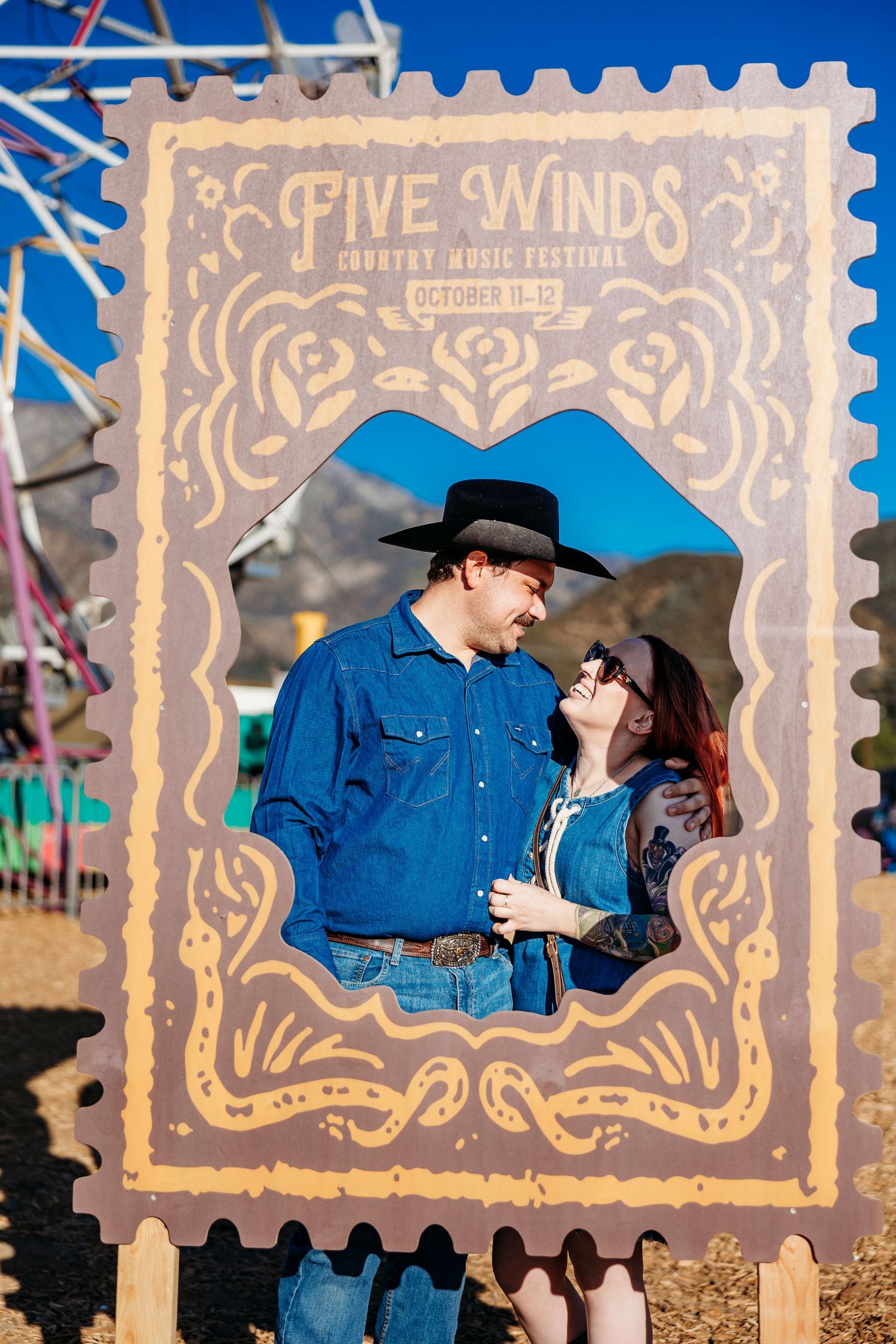 Couple poses behind decorative Five Winds festival frame in western attire.