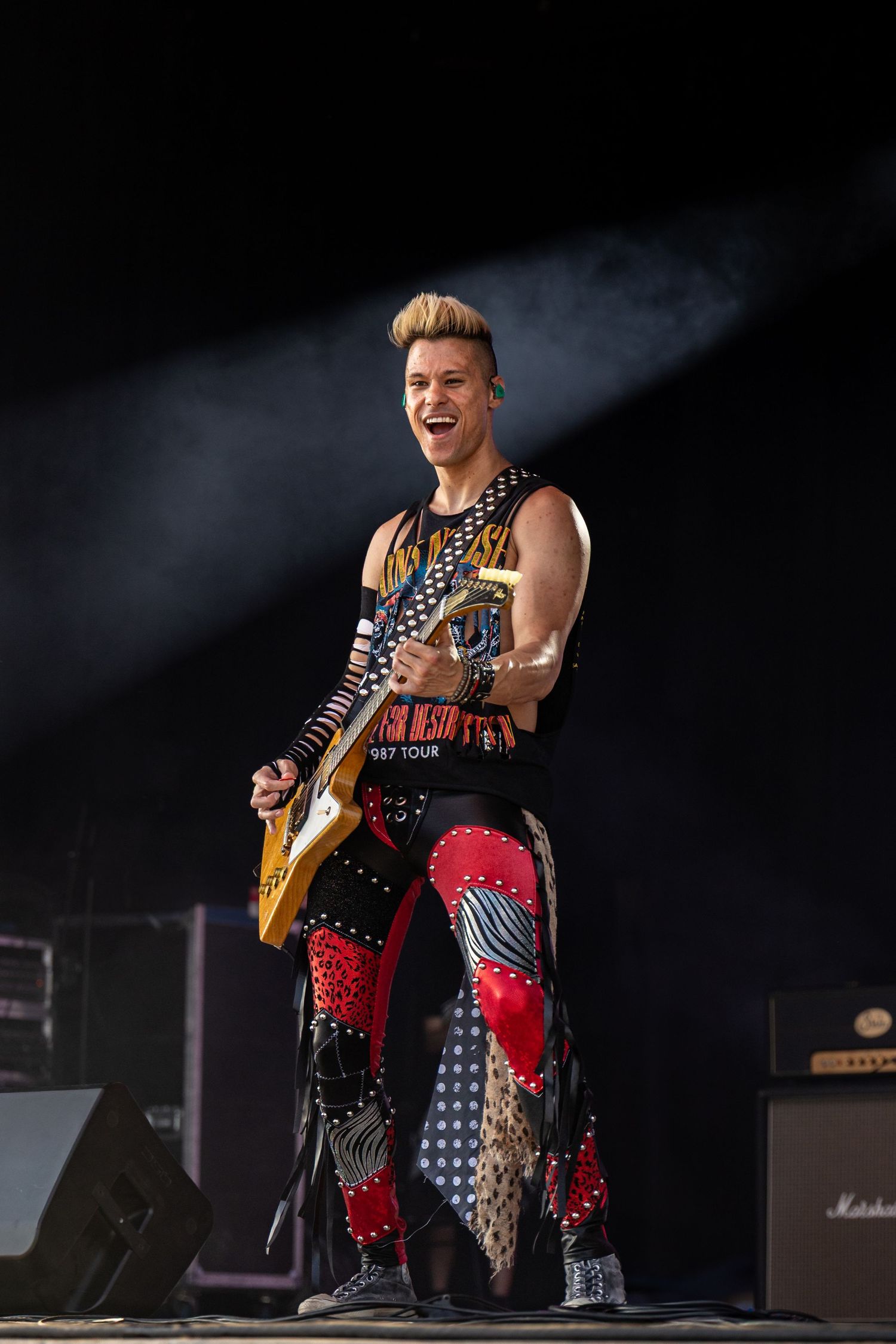 A guitarist performs on stage in patterned pants under dramatic lighting at a rock concert.