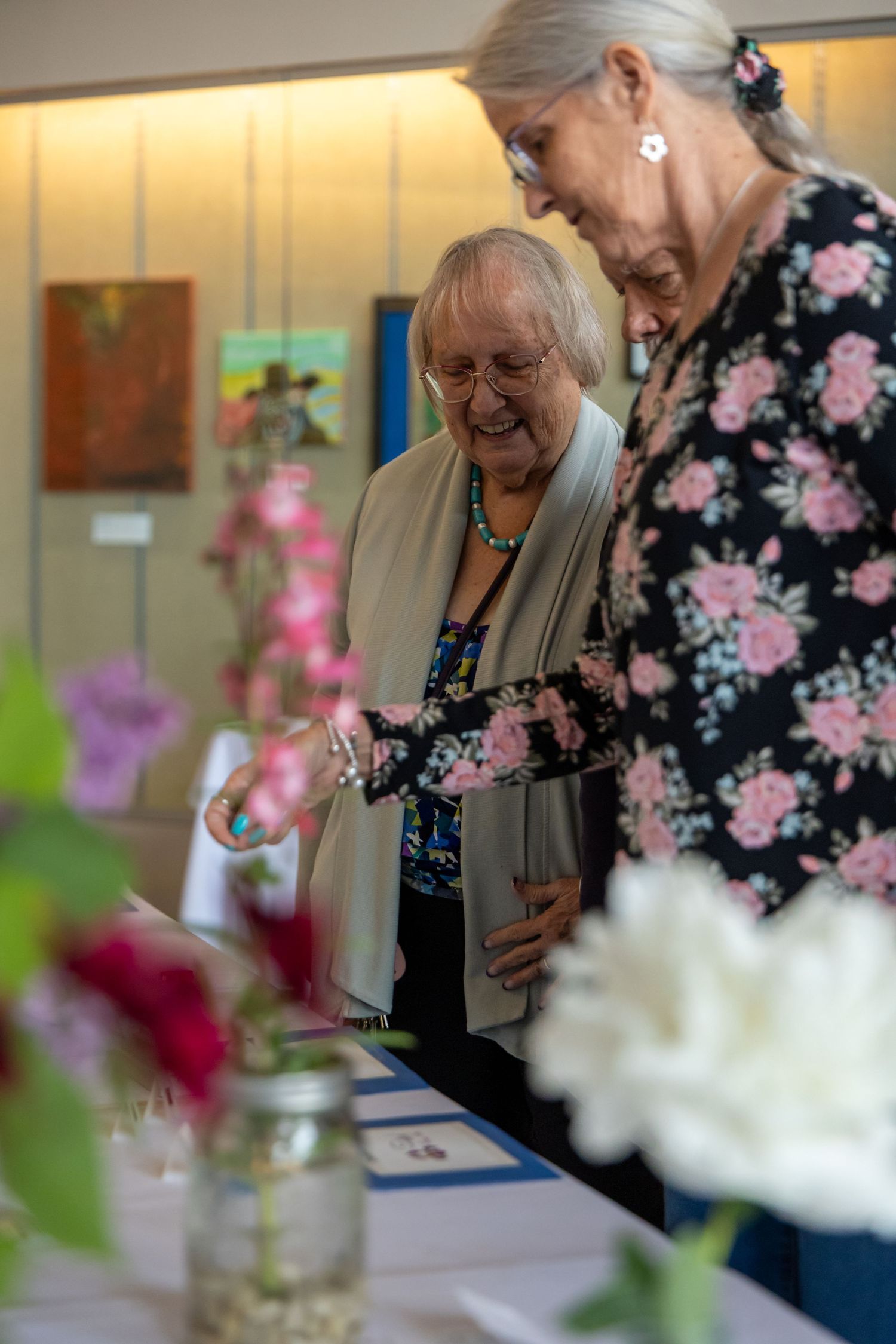 Two attendees share a joyful moment while looking at artwork displays in a bright gallery setting.