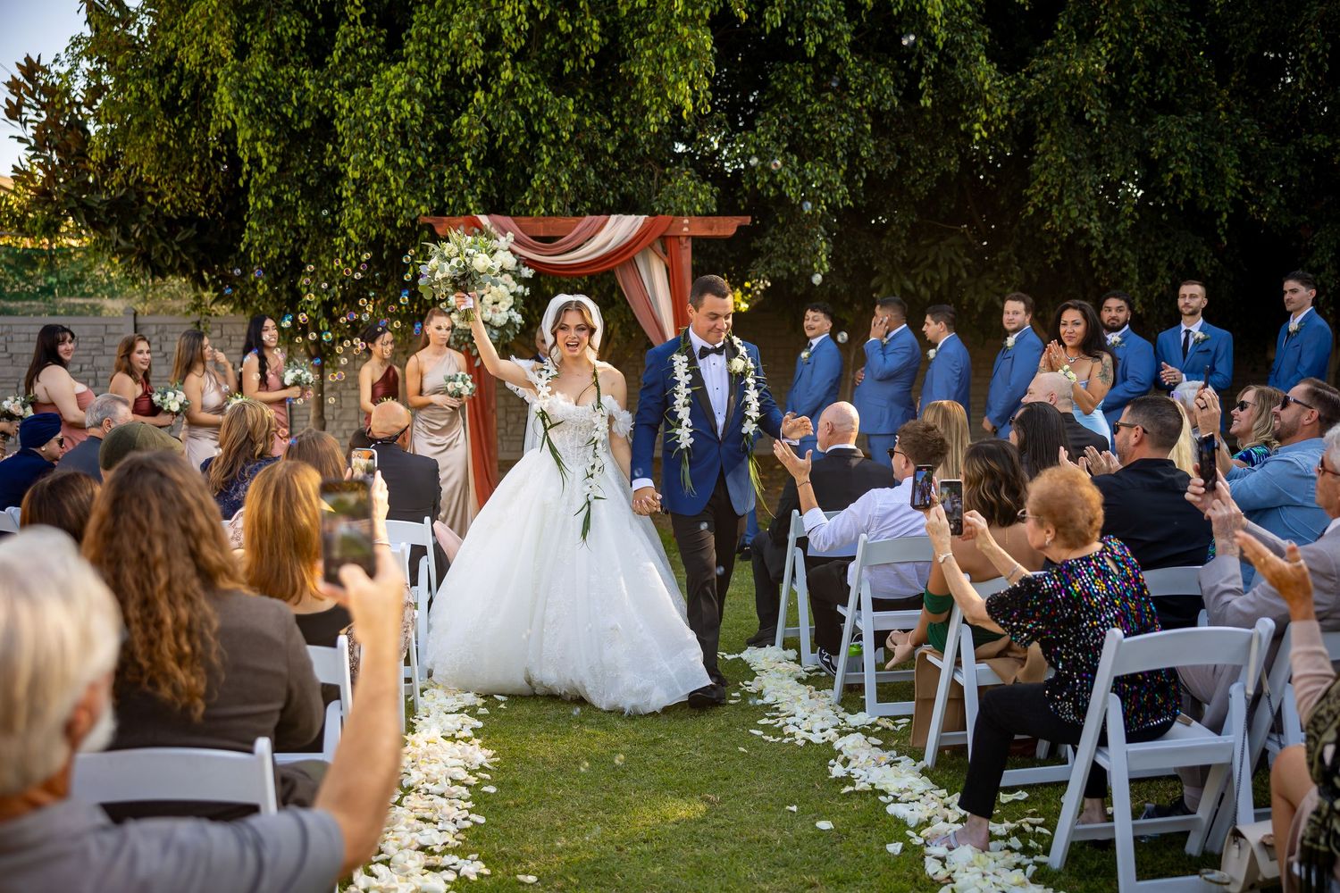Wedding ceremony taking place in a backyard garden setting with guests seated on white chairs.