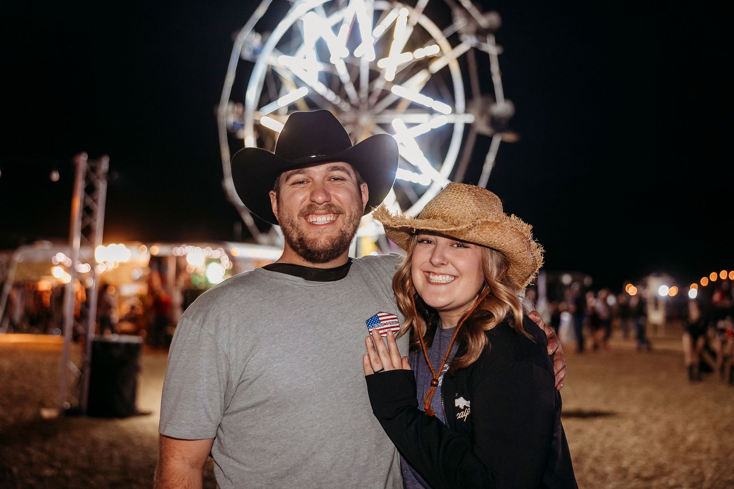A couple poses in front of an illuminated ferris wheel at night.