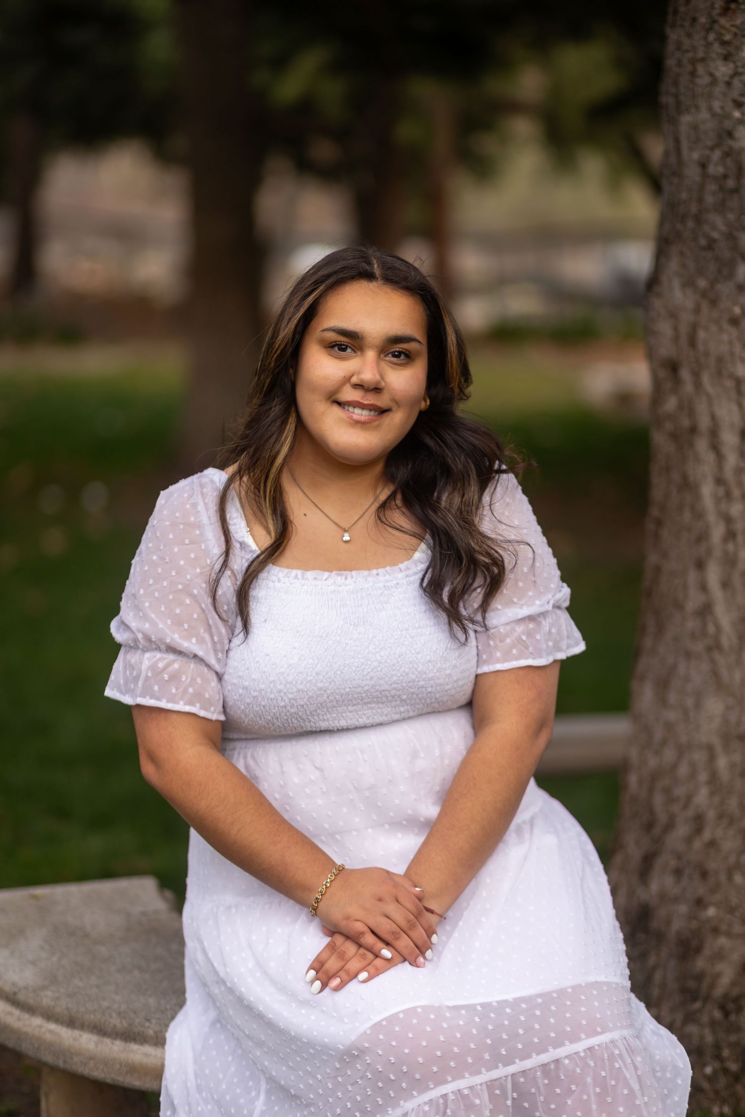Young graduate in white lace dress sitting outdoors on stone bench among trees in Oak Glen.