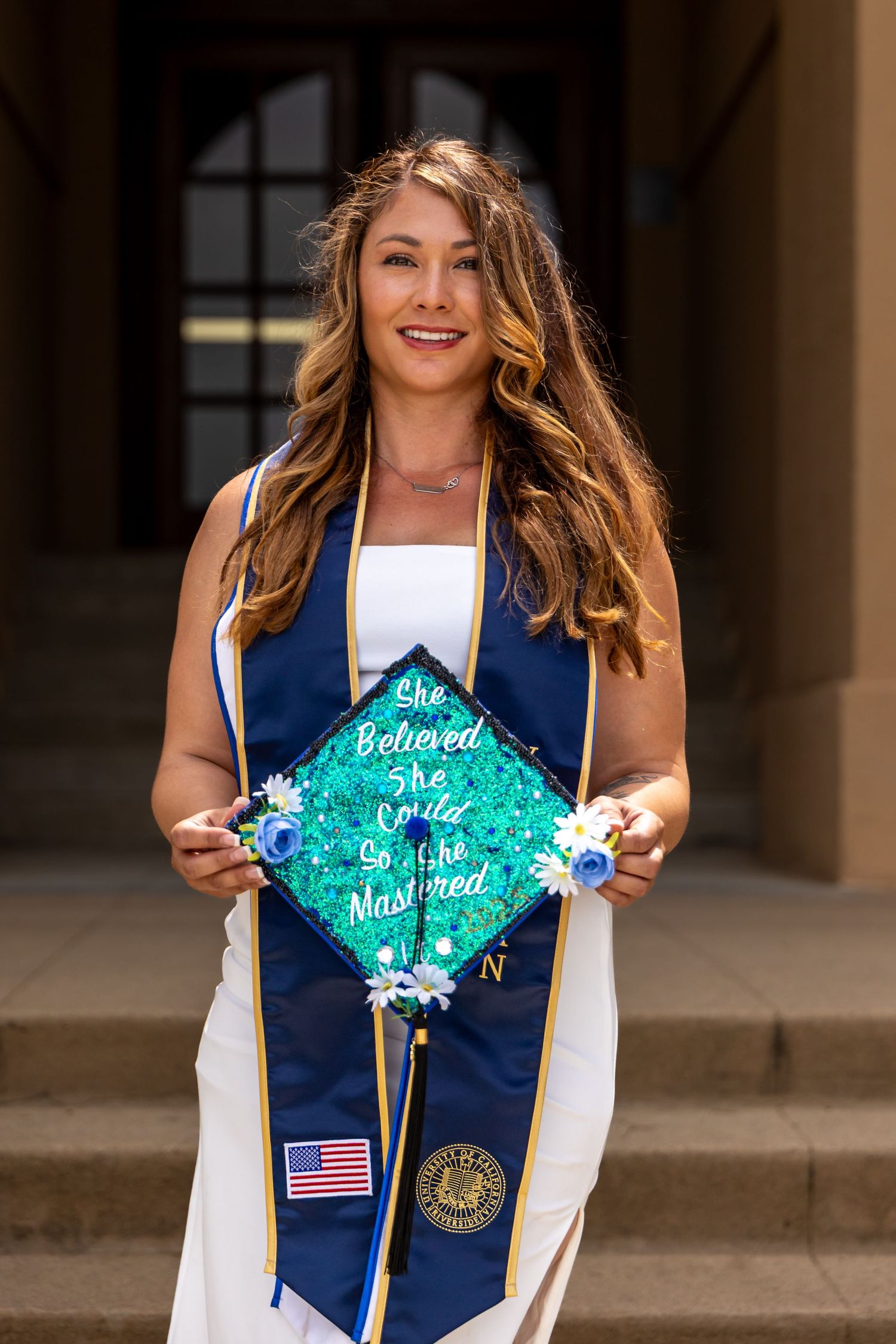 Graduate in white dress and blue stole displaying decorated graduation cap in Riverside.