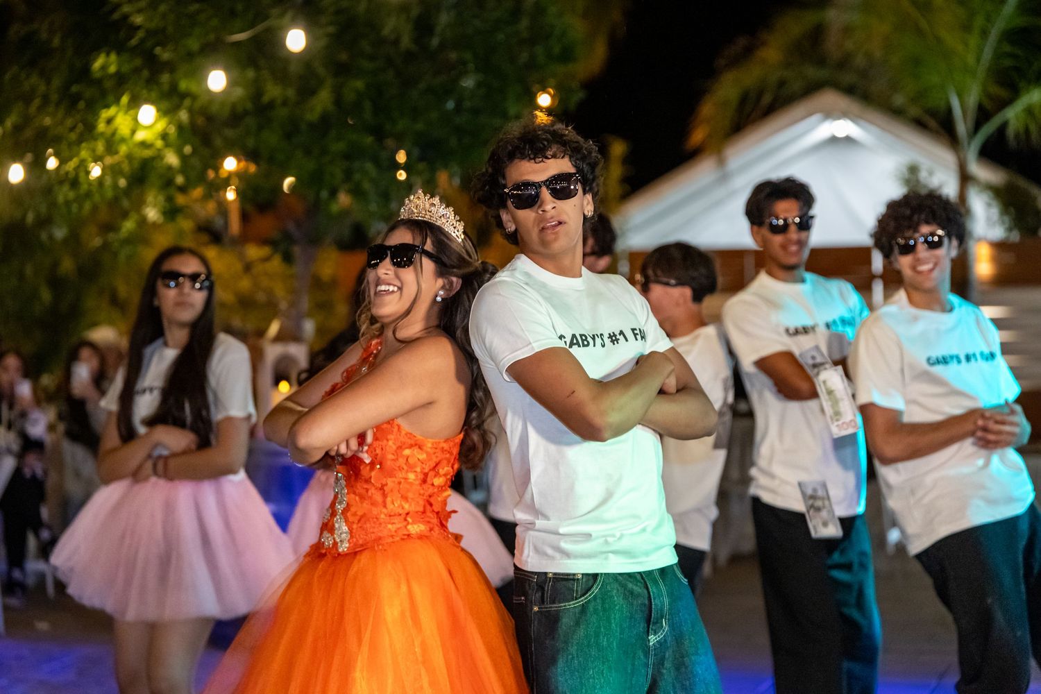 Party guests wearing sunglasses and tutus dance during an outdoor evening celebration.