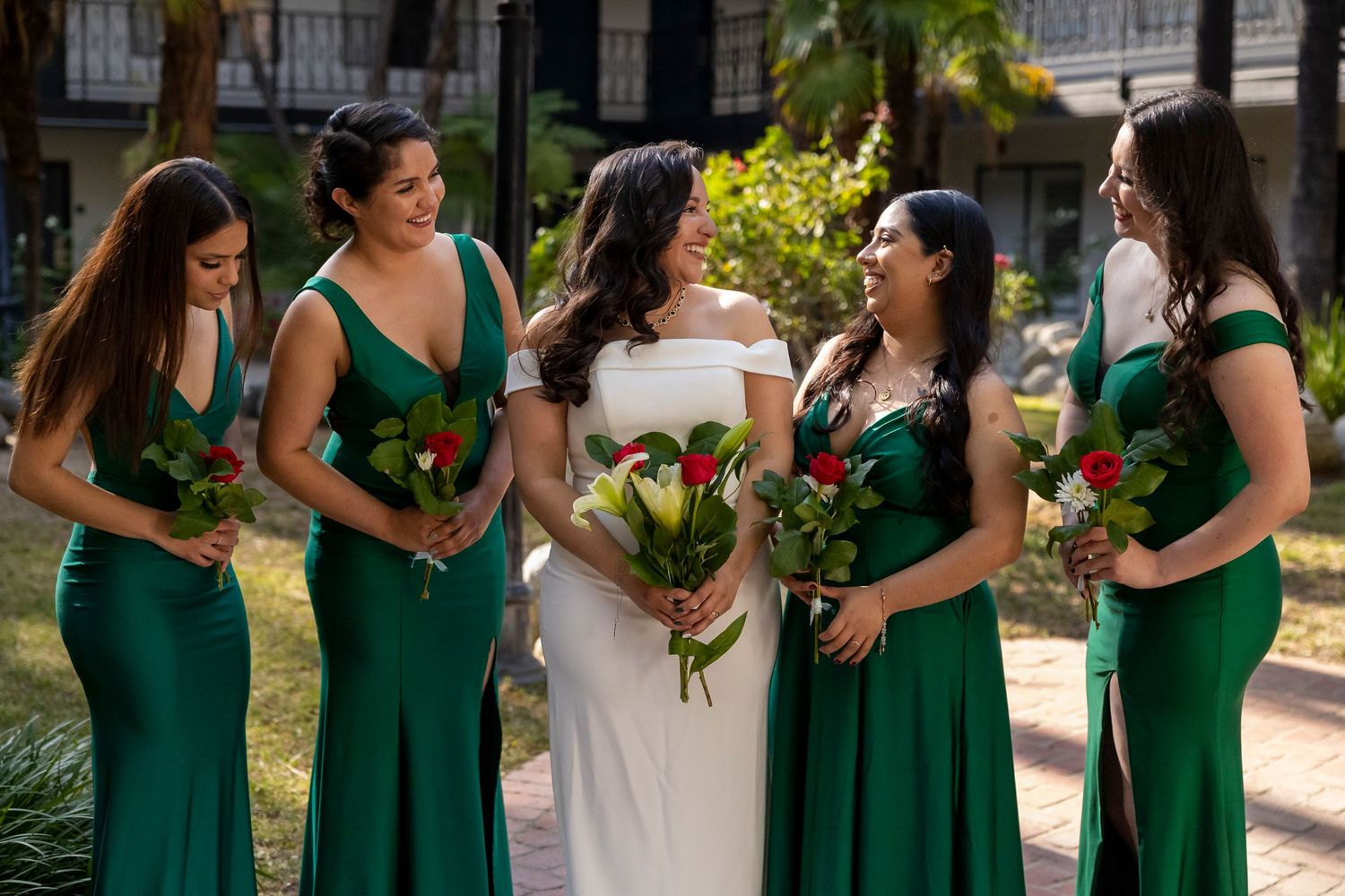 Wedding party pose outdoors with bridesmaids in emerald green dresses holding yellow bouquets.