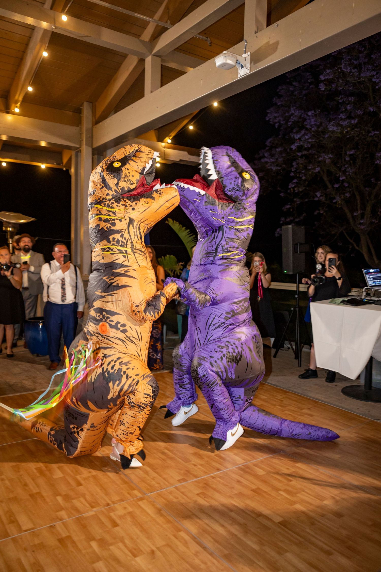 People in inflatable dinosaur costumes dancing on a wooden floor at a wedding reception.
