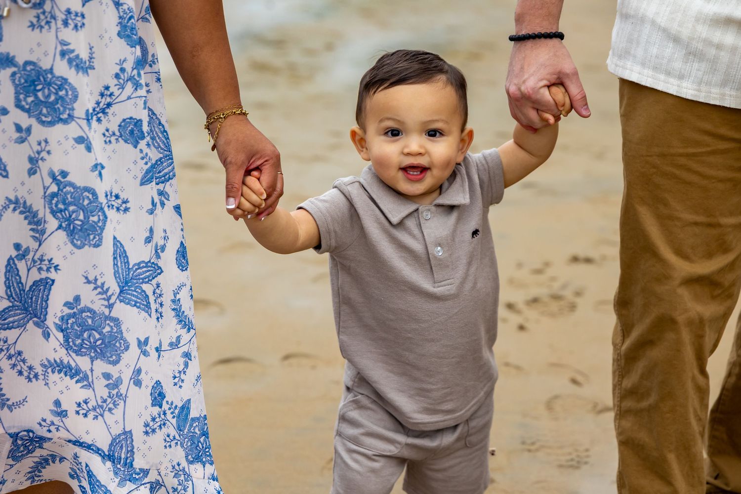 Small toddler in grey shirt walking on beach while holding hands with adults.