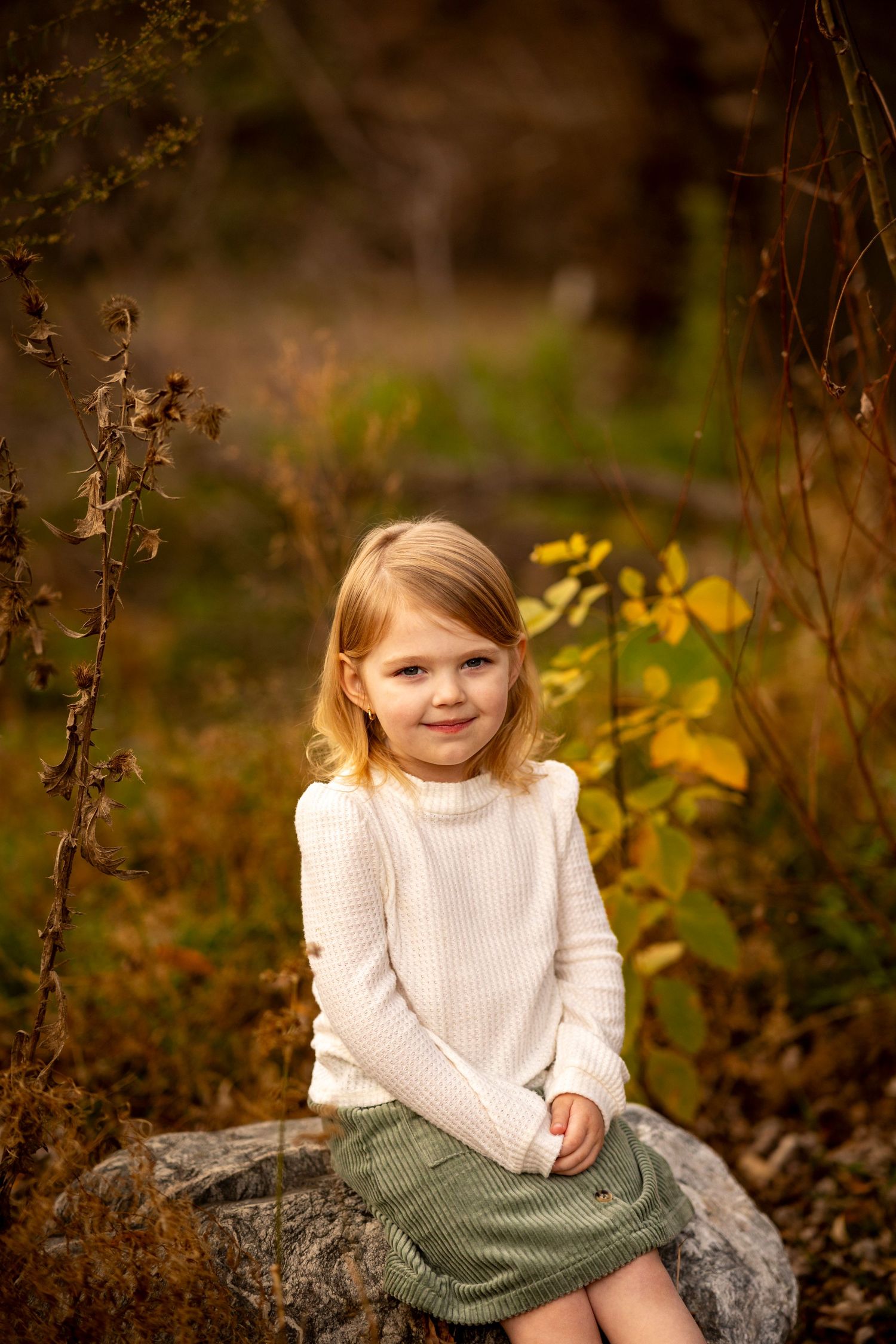 Child sitting on rocks outdoors wearing a white sweater and grey skirt in autumn scenery.