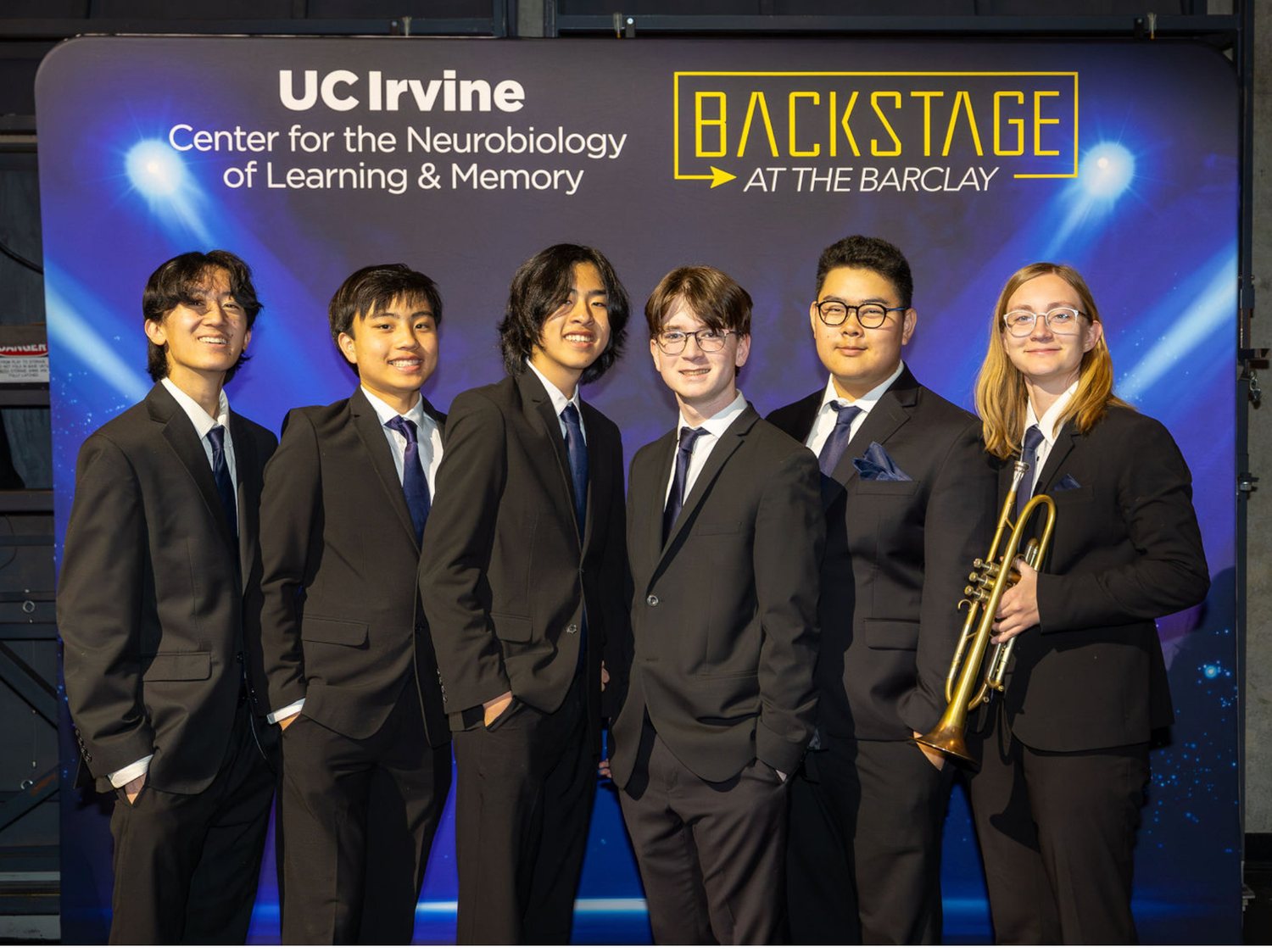 Musicians wearing formal black suits pose together on stage with a blue-lit backdrop for a performance at UC Irvine.