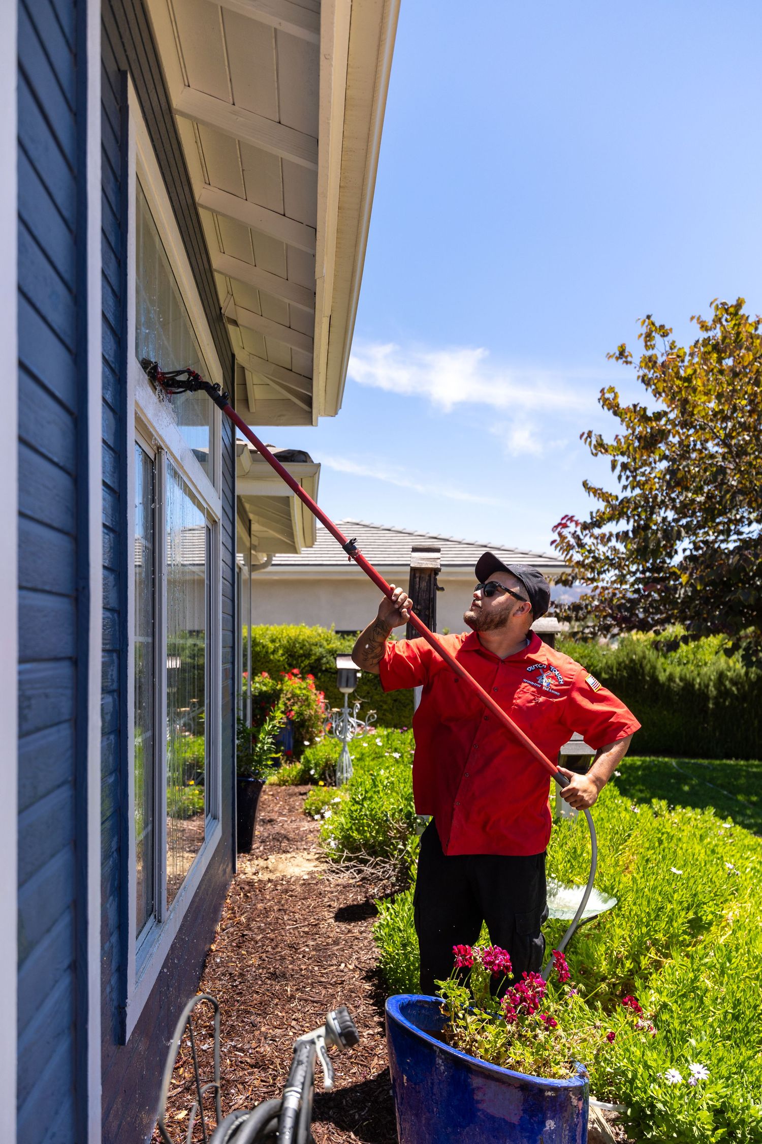 Someone washing exterior windows of a blue house with a long-handled brush while standing on a bucket in a garden.