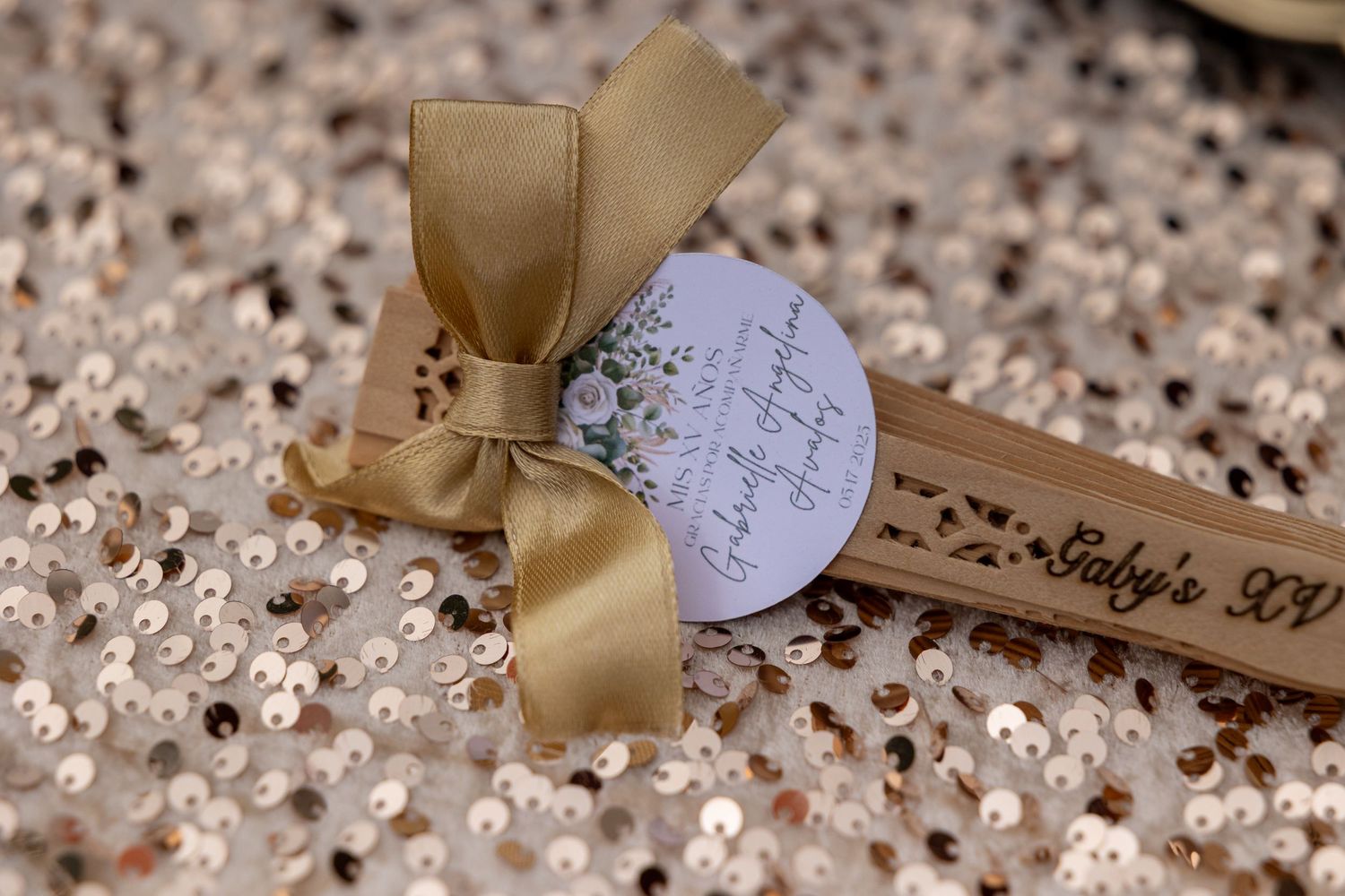 A wooden stirring stick with a gold bow and tag displayed on a sparkly sequined surface.