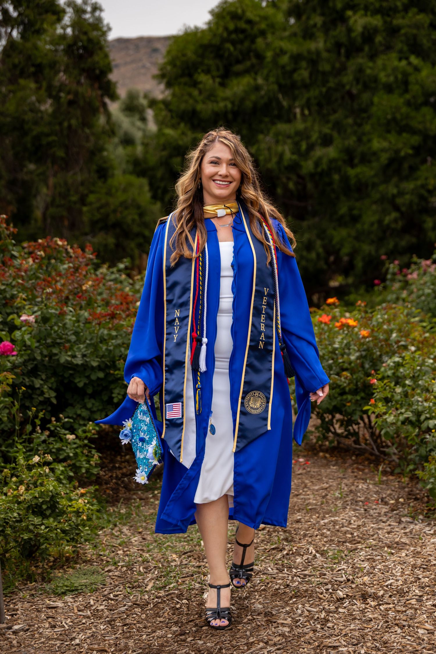 Graduate in blue cap and gown walking through a garden with colorful flowers in Riverside.
