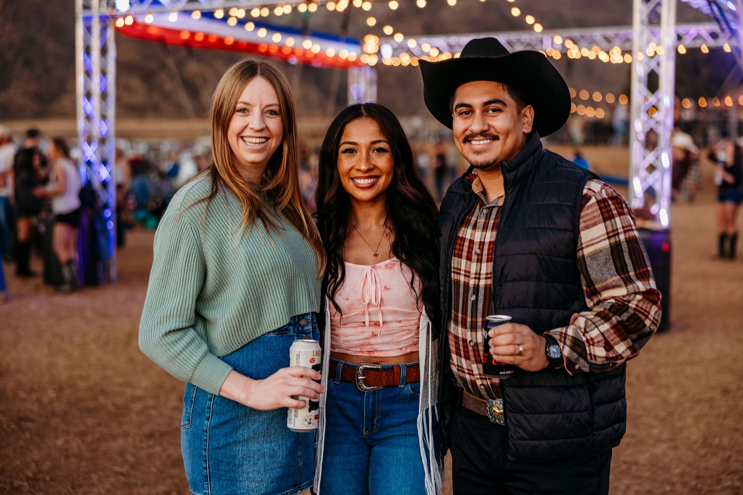 Three festival-goers pose together under string lights at an outdoor event.