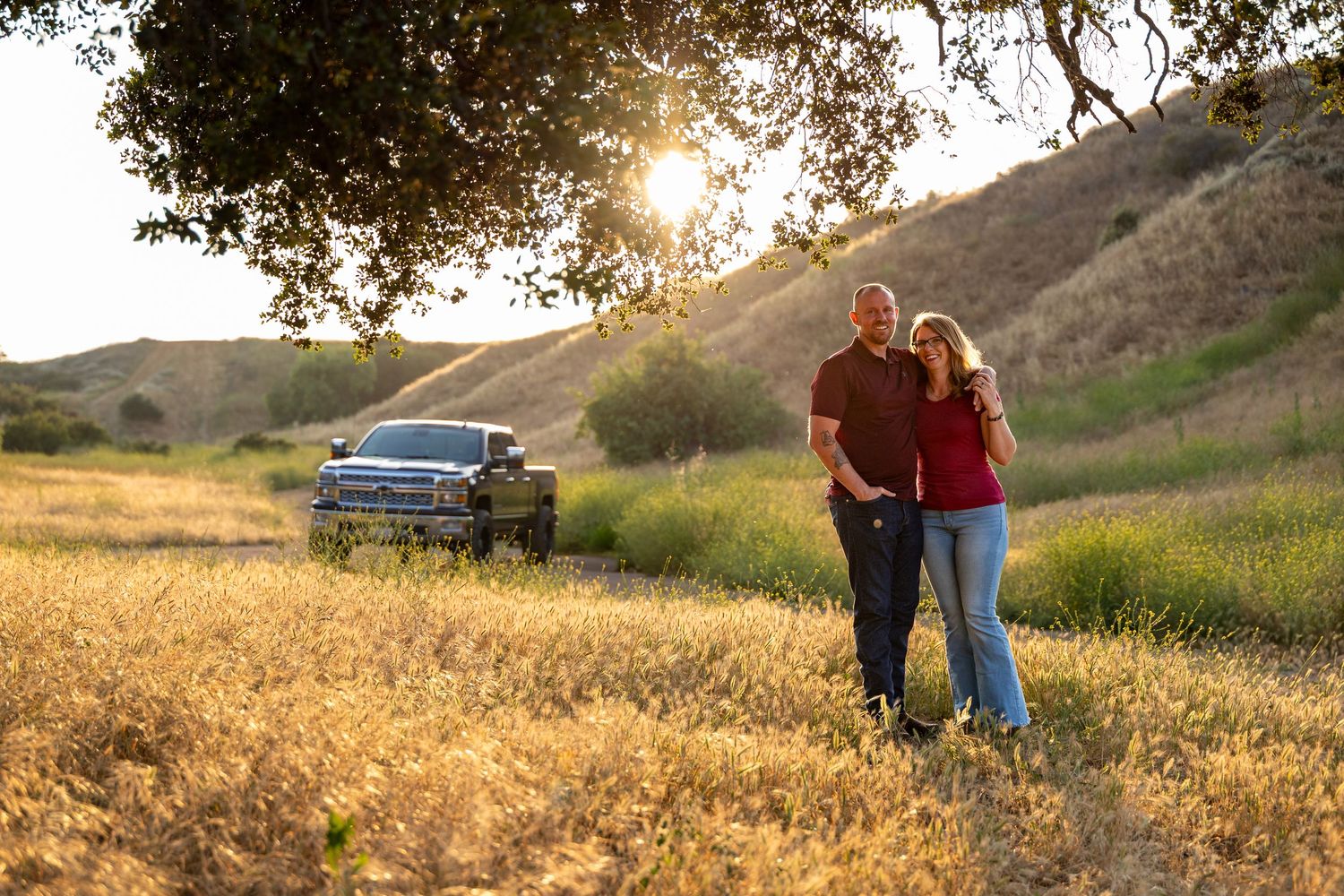Couple enjoying sunset in a golden meadow with their vintage car parked nearby.