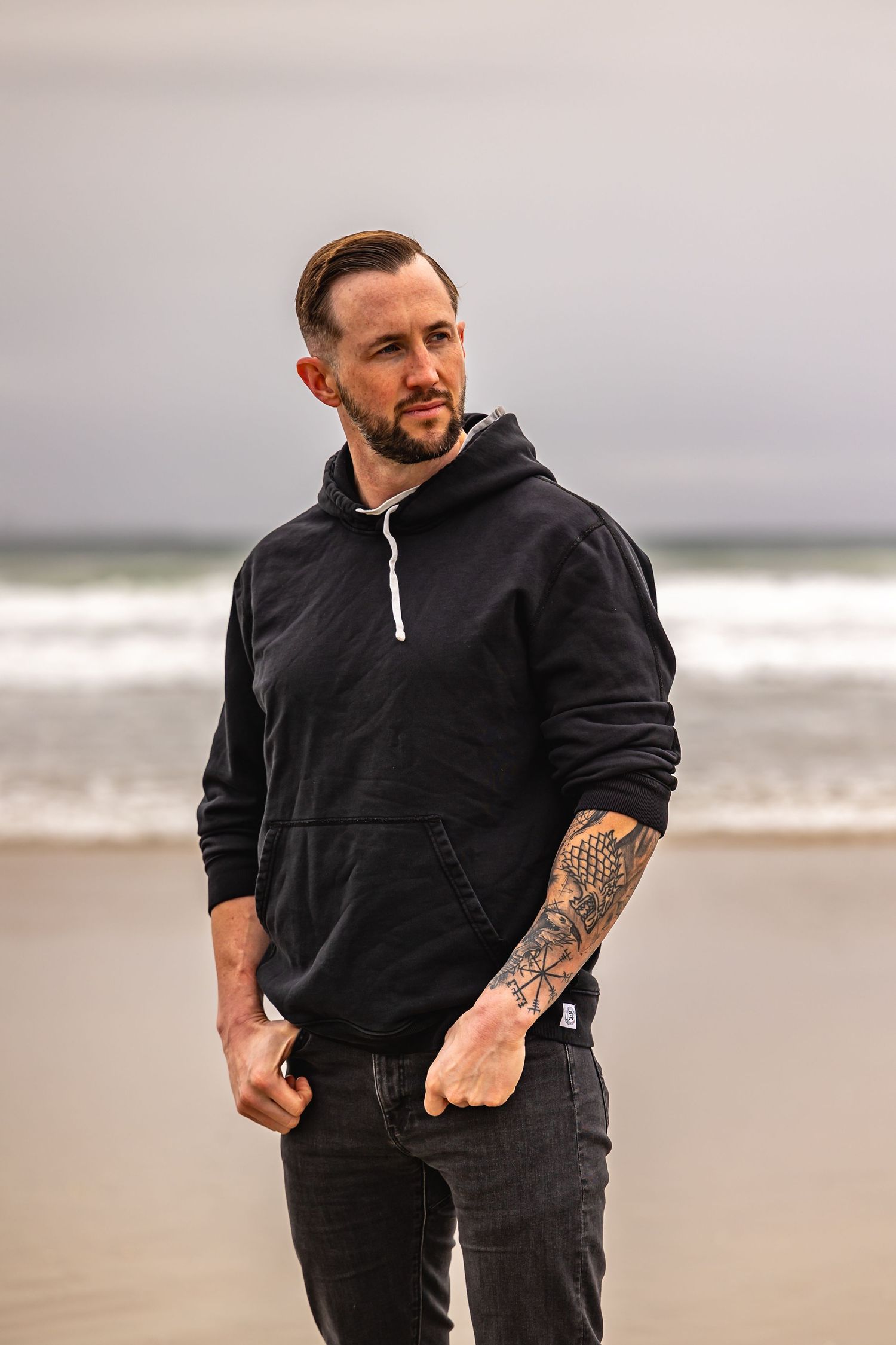Person in black sweatshirt walking on beach shows arm tattoos in overcast weather in Huntington Beach.