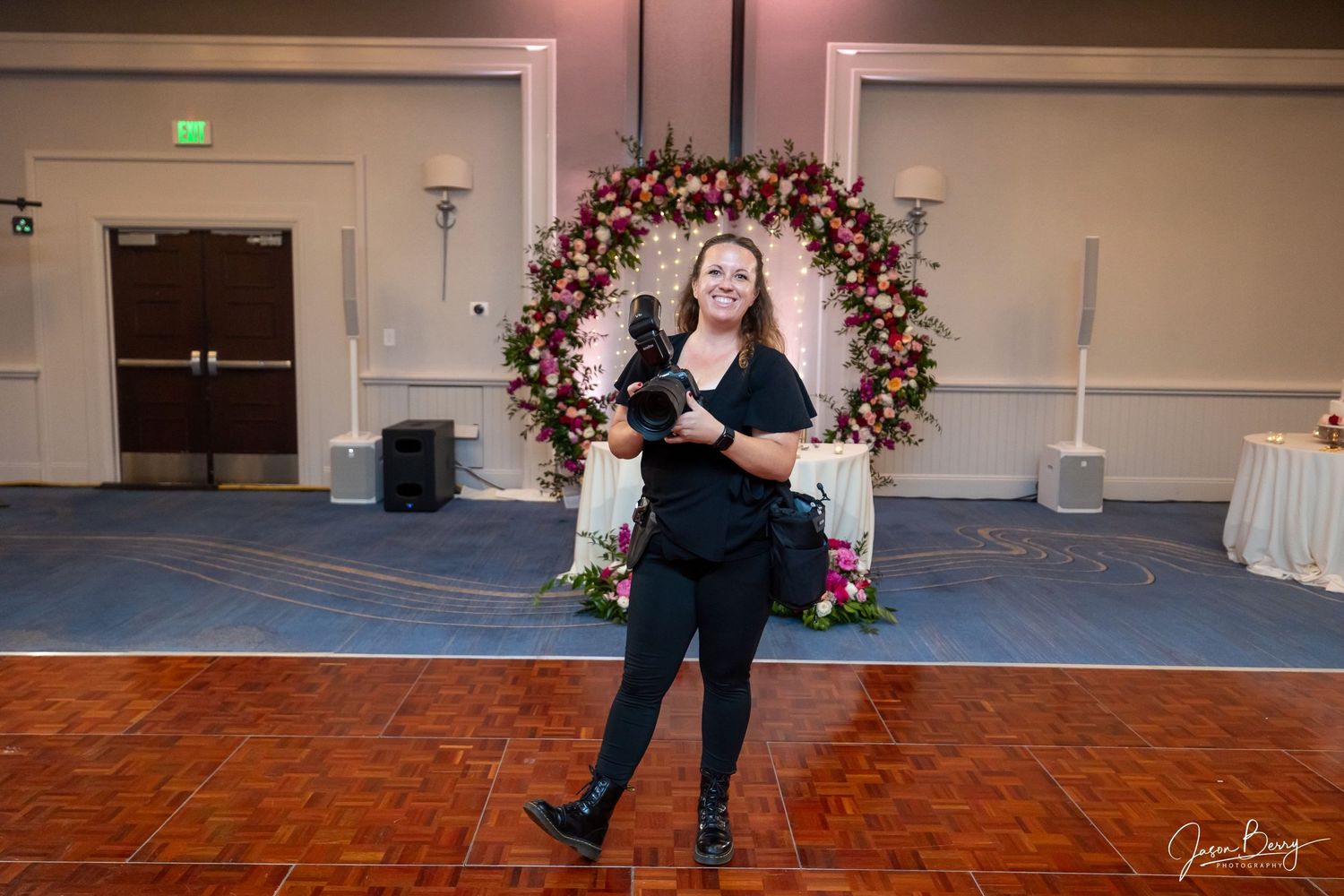 A figure in all black attire stands in front of a decorated floral arch at an event venue with a dance floor.