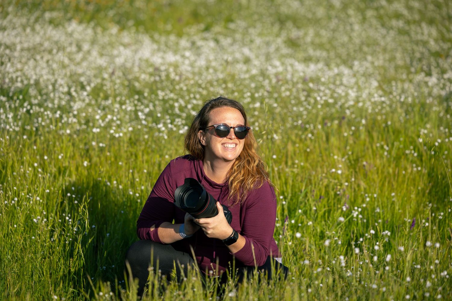 Person lying in field of white wildflowers smiling at camera in sunlight.